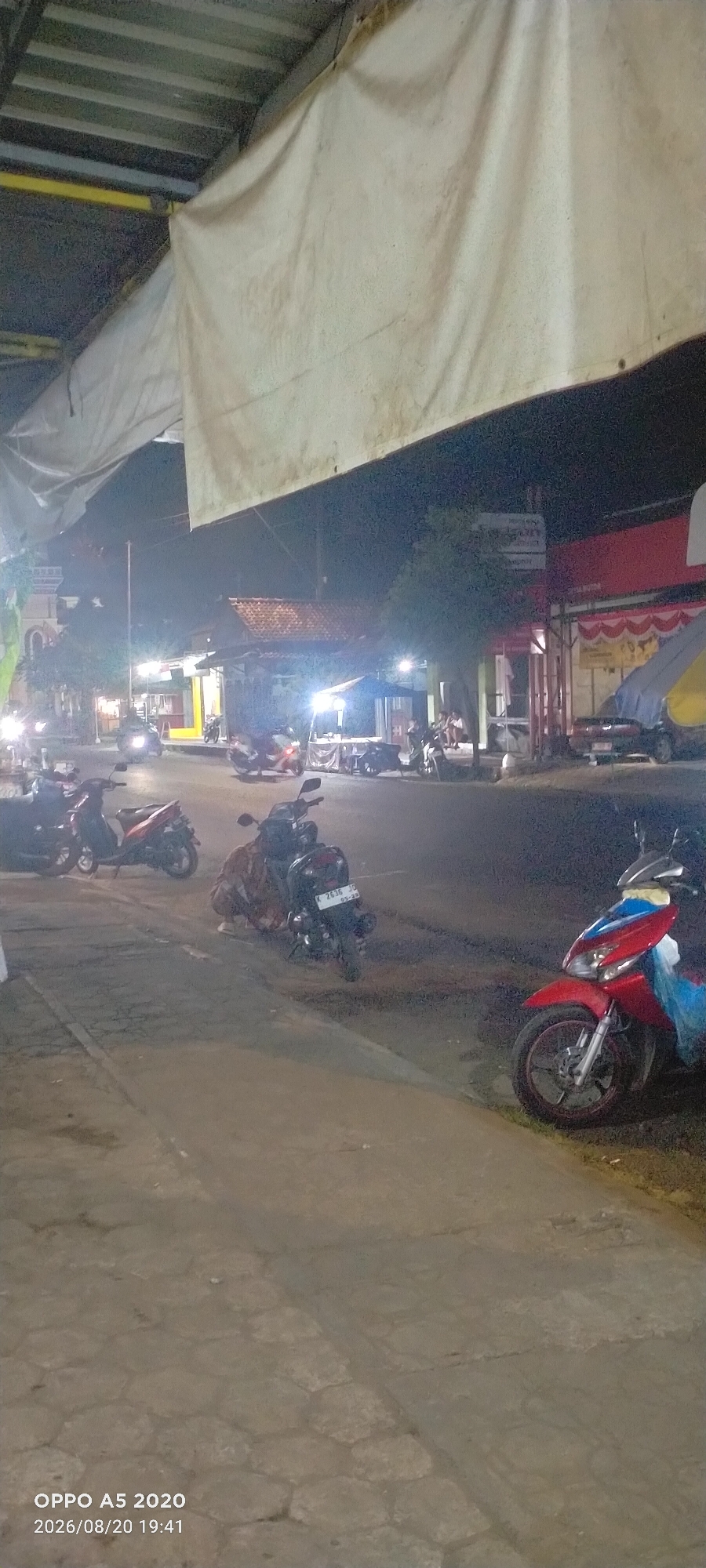 The image shows a nighttime street scene in Kedung, Indonesia. Several motorcycles are parked along the side of the road. Some are illuminated by bright lights from nearby buildings. In the background, there are commercial establishments, including what appears to be a food stall with tables and chairs, and a building with a red facade. The street is paved and there is a sidewalk. A large, off-white tarp is suspended overhead in the foreground, partially obscuring the view of the sky. The timestamp on the image indicates it was taken on August 20, 2026, at 19:41.