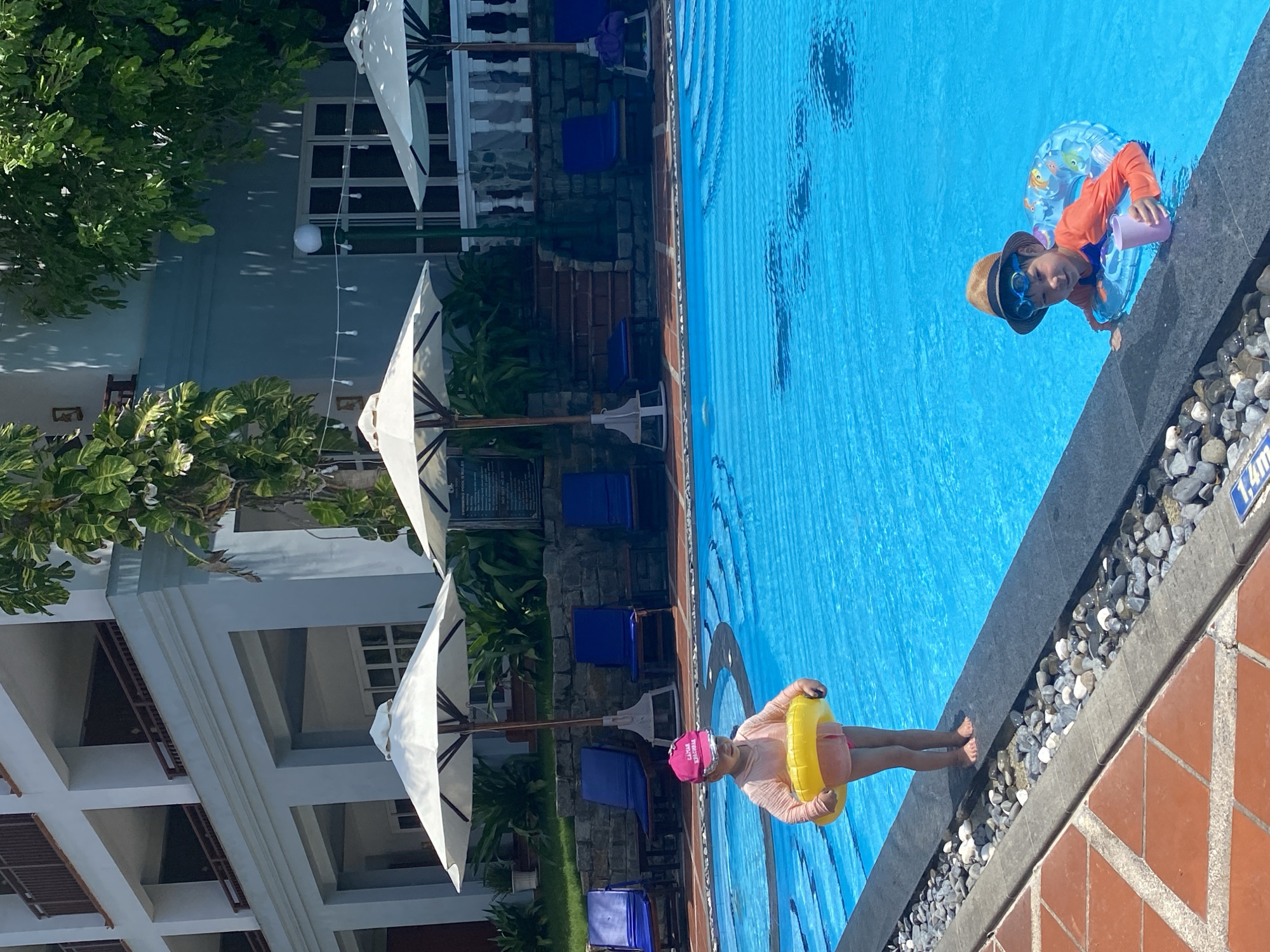 The image depicts an outdoor swimming pool scene at a resort in Phu Thuy Ward, Vietnam, under bright daylight.
In the foreground, a reddish-brown tiled patio area transitions to a dark grey pool coping, which features a decorative strip of smooth grey, white, and black pebbles. A partially visible blue and white sign on the pool coping indicates a depth of "1.4m".
Two young children are present at the pool. On the left, a child wearing a pink long-sleeved swim shirt, pink swim bottoms, and a pink baseball cap with "CAMIA" text stands barefoot on the pool coping, holding a yellow inflatable ring around their waist and looking down. On the right, another child, wearing an orange long-sleeved swim shirt, blue swimming goggles pushed up on a brown fedora-style hat, and a blue inflatable ring with fish patterns, is partially submerged in the pool, smiling towards the camera while holding a light purple plastic cup in their right hand, with their left arm resting on the pool edge.
In the mid-ground, behind the pool, three large white outdoor umbrellas with wooden poles provide shade over several blue lounge chairs with dark frames, some appearing to have towels draped over them. Lush green foliage, including potted plants and larger trees, is abundant. Stone steps lead upwards to different levels of the resort. A string of white globe lights is visible between architectural features.
In the background, a multi-story building with a light blue/grey facade features balconies with brown wooden railings. Additional green trees and foliage are visible around and behind the building, casting shadows. The scene is illuminated by strong sunlight.