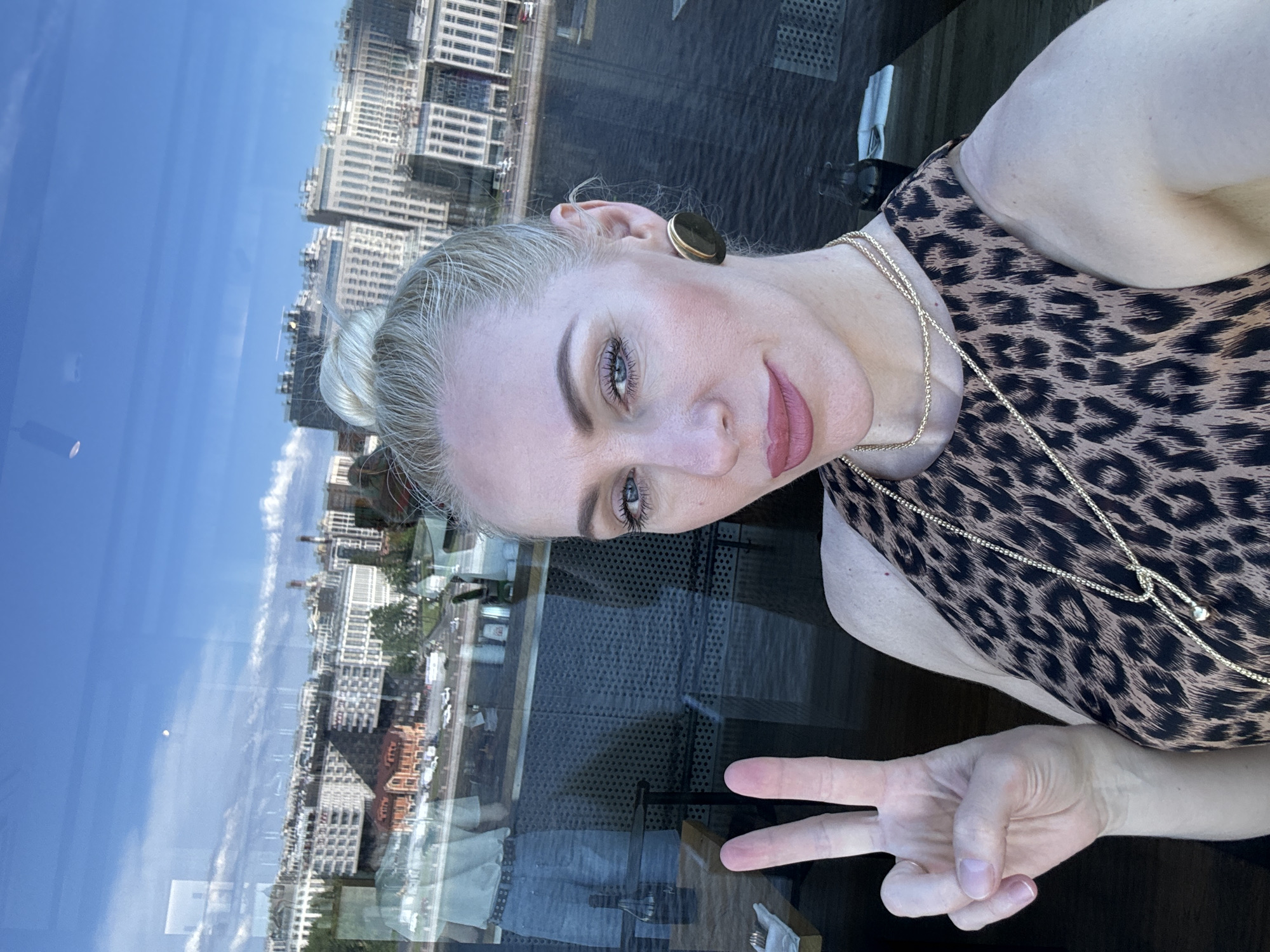 A blonde woman takes a selfie, looking directly at the camera with a slight smile and her left hand raised in a peace sign. Her hair is styled in a high bun, and she wears a leopard print sleeveless top, multiple gold-colored necklaces, and large gold disc earrings. Her makeup includes blue eyeshadow, eyeliner, and pink lipstick.
The image is captured through a large glass window, revealing a panoramic view of Saint Petersburg, Russia. A wide body of water, likely a river or canal, dominates the middle ground, bordered by a tree-lined street with several parked and moving vehicles. Beyond the waterfront, a dense cityscape extends, featuring numerous multi-story buildings of varied architectural styles, including some older, reddish-brown structures and many modern buildings with light facades and rectangular windows. The sky is bright blue with scattered white clouds; a faint white circular object, possibly the moon, is visible in the upper sky.
The glass window displays prominent reflections from the interior: a partial reflection of a person wearing a light shirt and light blue shorts or jeans, standing further back in the room. Other reflections include a dark bottle and a white folded napkin on a dark table, which partially obscures the water and lower buildings. The woman appears to be seated at this dark-surfaced table.