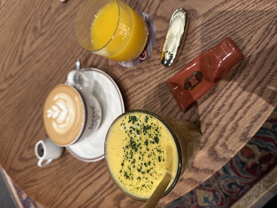 A wooden table displays a vibrant array of beverages and objects. A glass of orange juice and a latte with a milk design share space with a green-speckled yellow drink with a straw. Adjacent to the juices lie a golden, textured object and a small, red package. The scene is well-lit, emphasizing the varied textures and colors on the table. A rug or patterned surface peeks into the lower right corner.