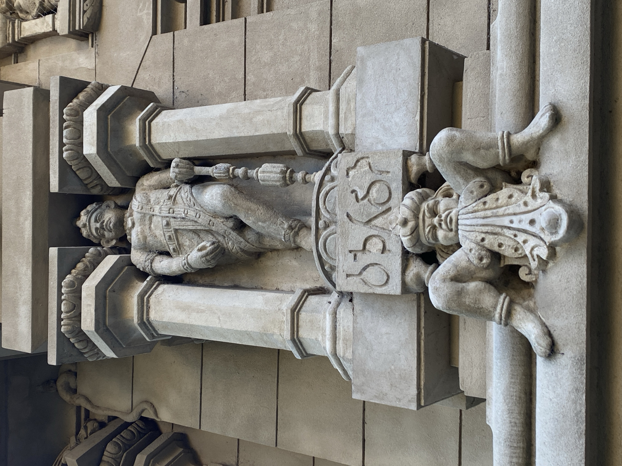 The image shows a stone sculpture of two figures, one a male figure in dress, the other a figure with legs and feet sticking out from a base, possibly a grotesque or allegorical figure. The sculpture is part of a building facade in Buenos Aires, Argentina. The building is likely from the early 20th century, based on the architectural style. The sculpture is mounted horizontally on the side of a building. The light suggests it is daytime.