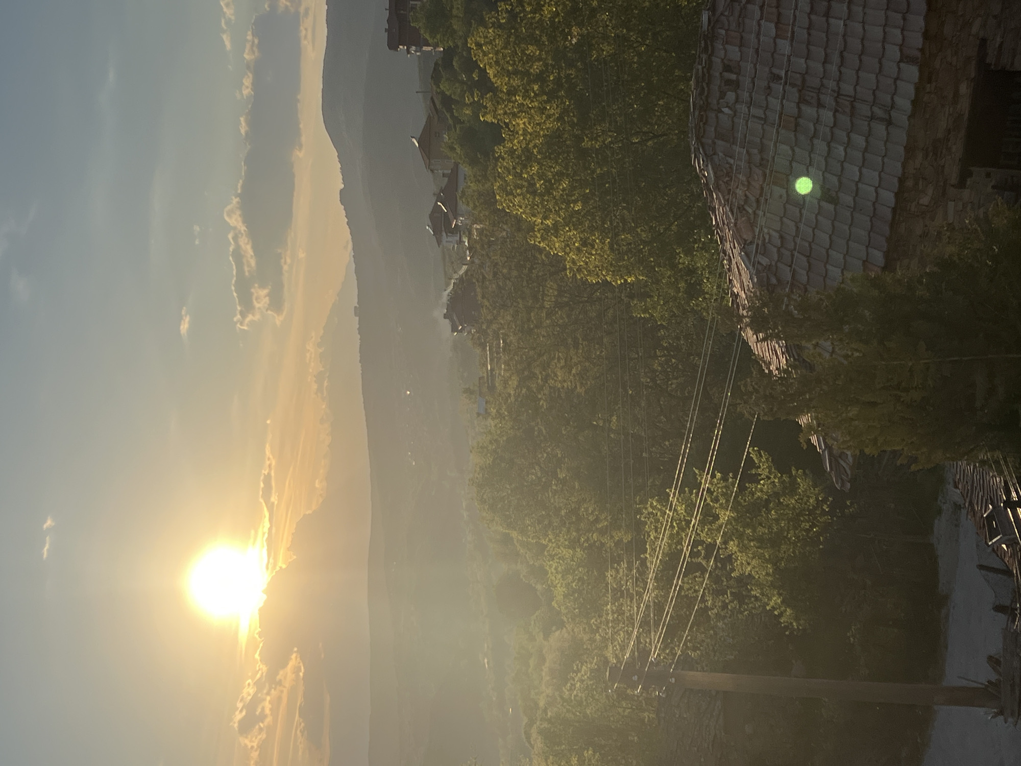 The image depicts a landscape at sunrise or sunset, likely in Kribul, Bulgaria, based on the presence of mountainous terrain and village architecture. The sun is prominently positioned on the left side of the frame, partially obscured by clouds, casting a bright, warm light across the scene.
A mountain range is visible in the background, partially obscured by haze or mist. Below the mountains, a village or town is discernible, with buildings clustered together. A wooden roof is in the foreground on the right side of the scene. Power lines run through the midground, and the presence of trees suggests a partly forested area. The overall impression is of a rural or semi-rural environment.