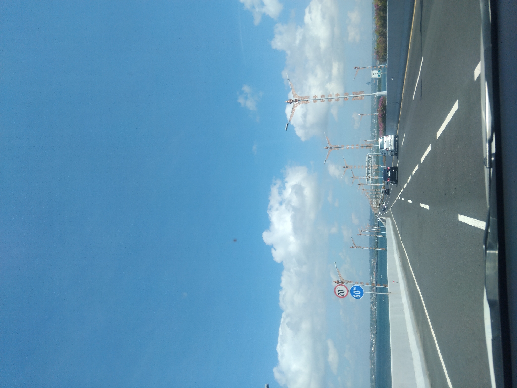 This image captures a view from inside a vehicle traveling on the Bali Mandara Toll Road over Benoa Bay in Kuta Selatan, Indonesia. The foreground displays the top of the vehicle's dashboard and a partial windshield wiper blade. The primary subject is a multi-lane asphalt highway, with two lanes clearly visible in the direction of travel, delineated by white dashed lines. A low, white concrete barrier separates these lanes from oncoming traffic to the right, and an identical barrier defines the left edge of the road, bordering the blue water of Benoa Bay.
On the left side of the road, two circular speed limit signs are mounted vertically on a pole: an upper red-bordered sign indicating "80 KM" and a lower blue sign indicating "60 KM." A series of unique streetlights, characterized by tall, ornate, copper-toned structures with multiple radiating arms, line both sides of the highway, extending into the distant perspective.
Several vehicles are visible ahead, traveling in the same direction, including a dark sedan and a silver minivan. Further along the right side of the road, sections of the bridge structure are adorned with bursts of pink and purple flowering plants. A small, partially obscured green road sign with white lettering, showing the number "600," is visible on the right in the distance. The upper two-thirds of the image are dominated by a clear, bright blue sky with scattered white cumulus clouds, primarily concentrated above the horizon line. Two distinct, dark, circular smudges are present in the upper sky, indicative of lens debris. The scene is illuminated by bright daylight.