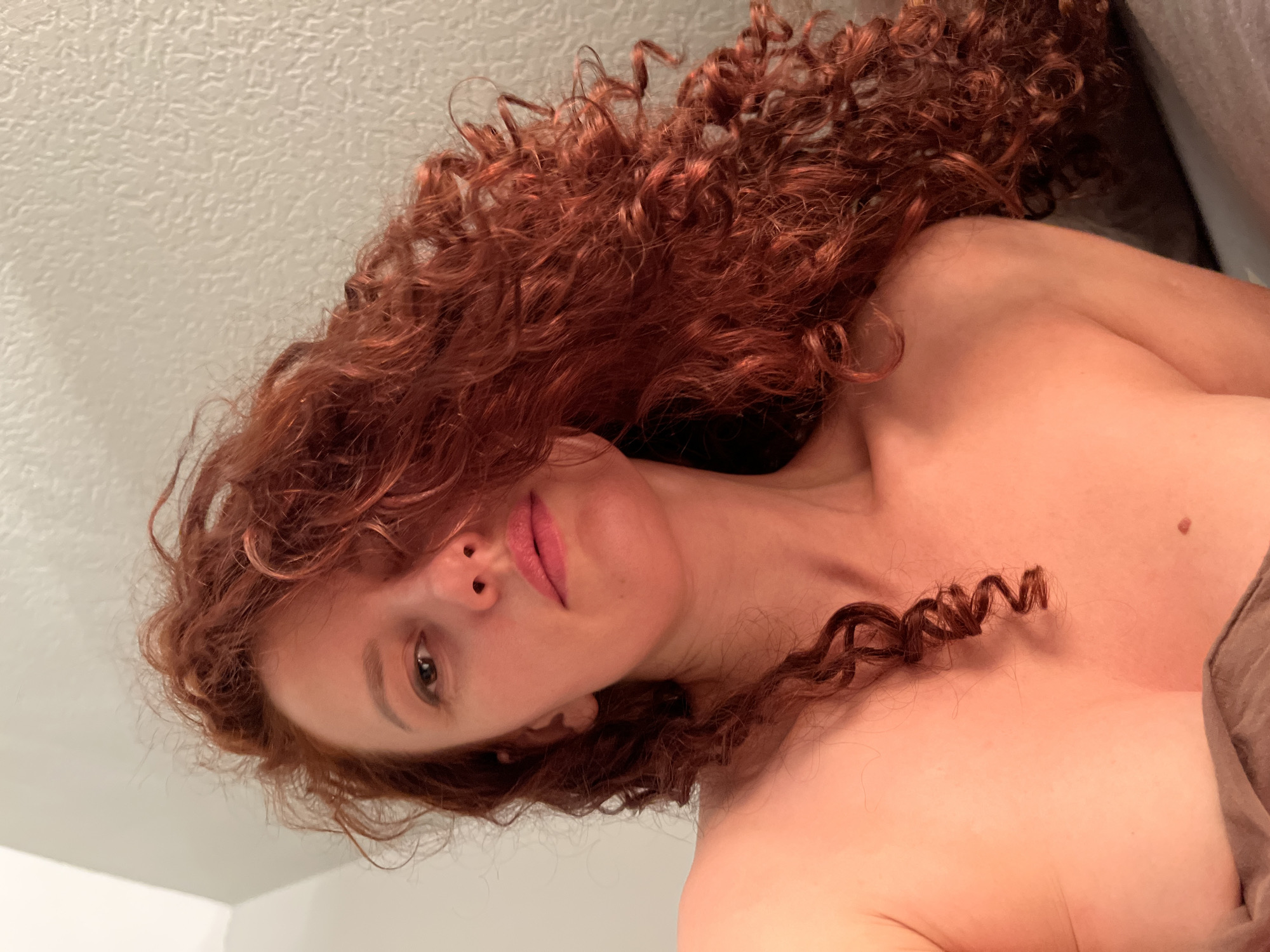 A woman with long, red curly hair is lying down. Her face is visible, and she is looking at the camera. A mole is visible on her chest. The background is a white wall. The image does not contain information that would identify Saint Petersburg, Russia.