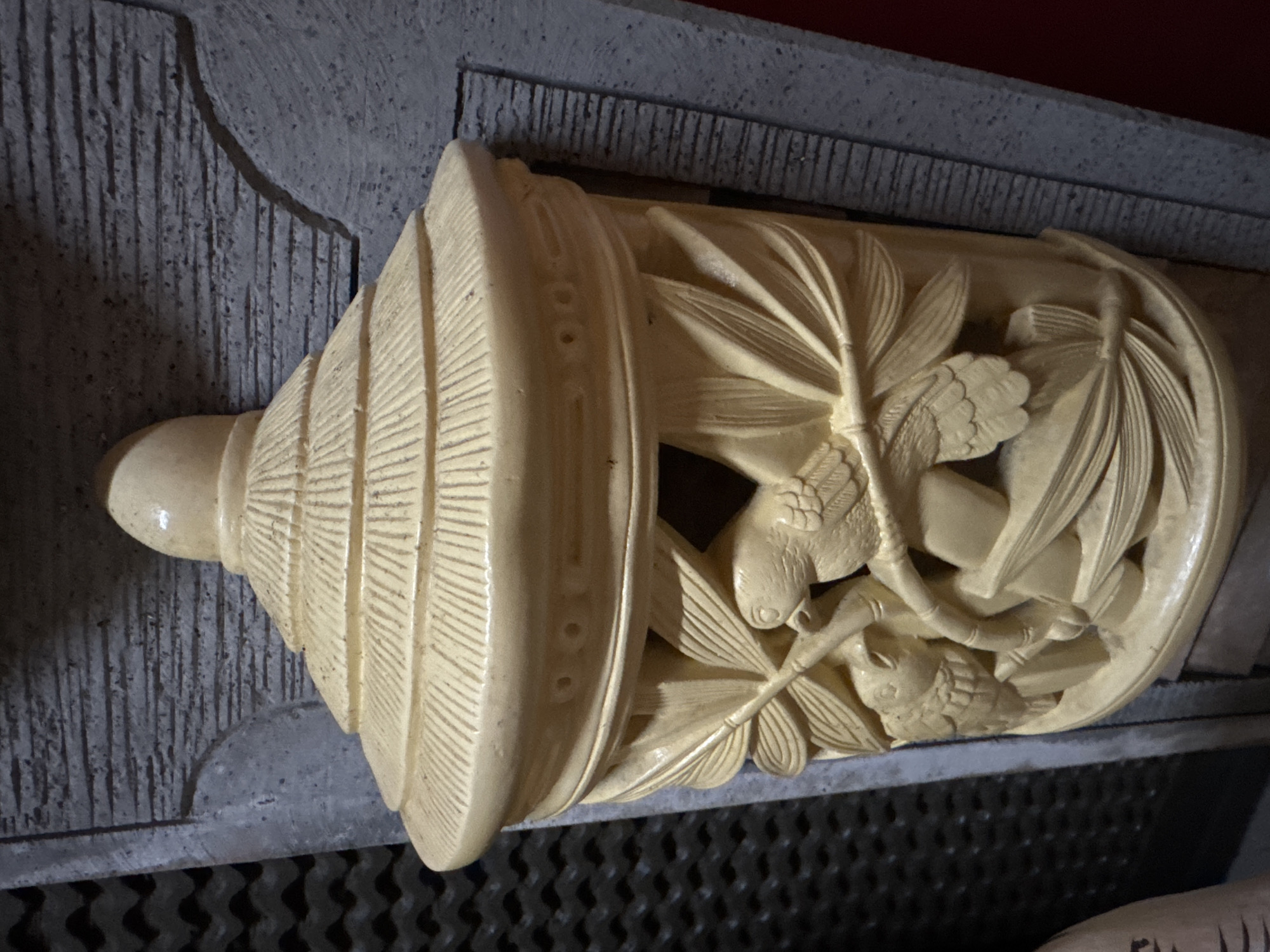 This media file showcases an intricately carved, light-colored object, possibly made of ivory or a similar material, featuring detailed depictions of birds and foliage. The object has a cylindrical body with a tapered end and is adorned with fine lines suggesting a ribbed texture. The carving on the main body portrays at least two birds perched on branches, surrounded by stylized leaves that resemble palm fronds or bamboo. The craftsmanship is detailed, with individual feathers and facial features on the birds clearly rendered. The object is placed on a textured, dark gray surface, possibly stone, which is set against a darker, perforated background. The lighting suggests an indoor environment, casting subtle shadows that highlight the relief of the carving. The orientation of the image is rotated, with the tapered end of the object pointing to the left. The overall aesthetic is one of delicate artistry and natural motifs. The location context of Kuta, Indonesia, suggests this might be a souvenir or decorative item originating from the region. There is no visible text in the image.
