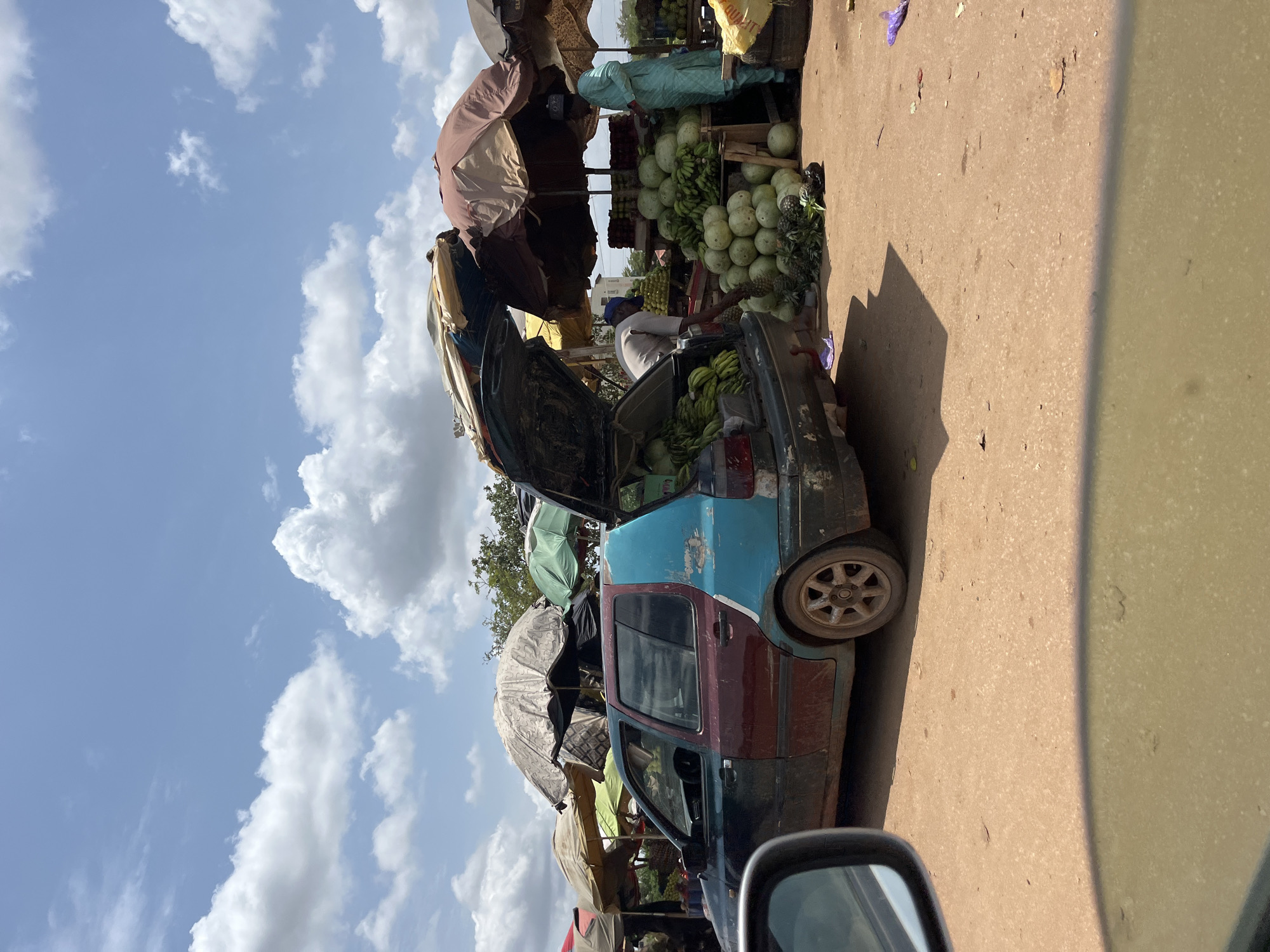In Galadimawa Village, Nigeria, a roadside market scene unfolds under a partly cloudy sky. The primary focus is a heavily used, multi-colored hatchback car parked on a dusty, unpaved roadside. The car's trunk is open, filled with bananas, suggesting it's being used as part of the market setup. Nearby, vendors stand under various sun umbrellas, selling fruits, including watermelons. The overall impression is of an informal, bustling marketplace.