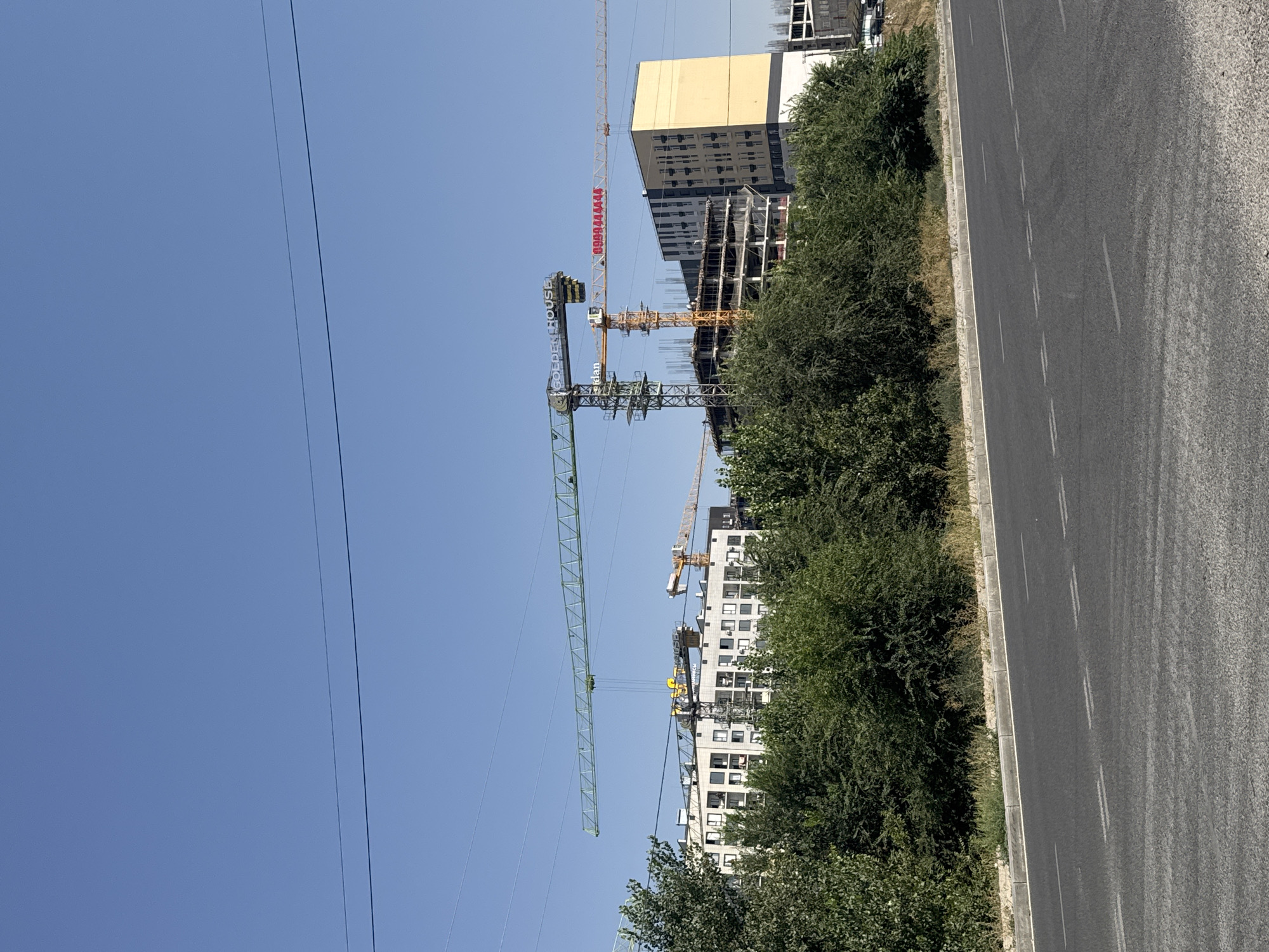 A bright, clear blue sky is visible above a construction site in Bishkek, Kyrgyzstan. Several cranes, including two large yellow and green ones, are actively engaged in construction. One crane has the text "GOLDEN HOUSE" and a phone number, "0909444444", printed on it. In the background, a multi-story building with a yellow facade is under construction, alongside other buildings and residential complexes. Lush green trees and bushes line the foreground, obscuring a portion of the construction site from view. A paved road with white lane markings runs across the bottom of the image, suggesting a public thoroughfare. The scene is well-lit, indicating it is daytime. There are no people visible in the image.