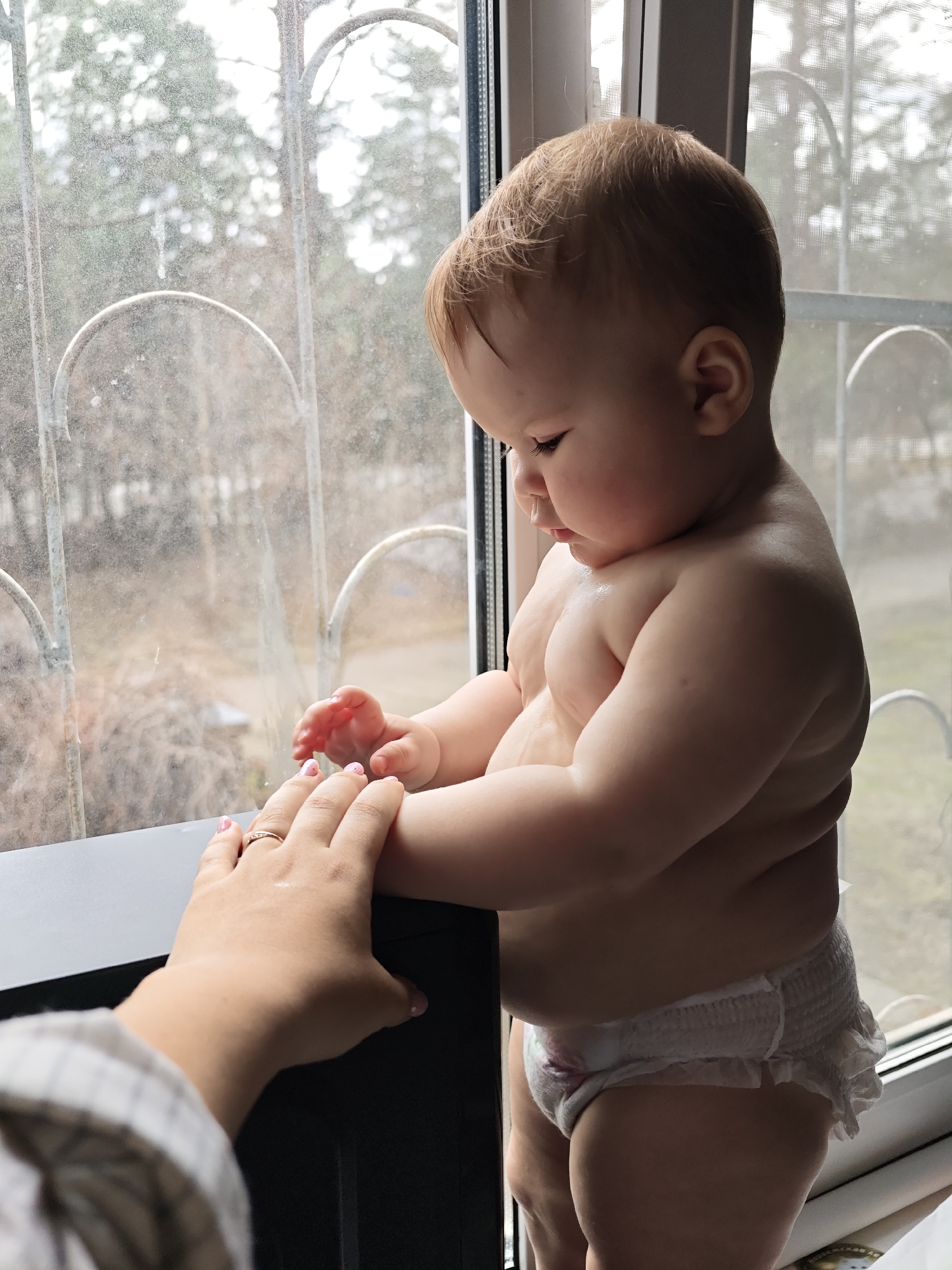 A baby wearing a diaper is standing near a window, holding onto the windowsill. The baby is turned slightly to the right and looking down. An adult's hand with painted nails is visible on the left, also holding onto the windowsill near the baby's hand. The window is made of metal and appears to be dirty. The background through the window shows blurred trees. Based on the metal window and architecture, the scene may be set in a city like Chaikovsky, Russia.