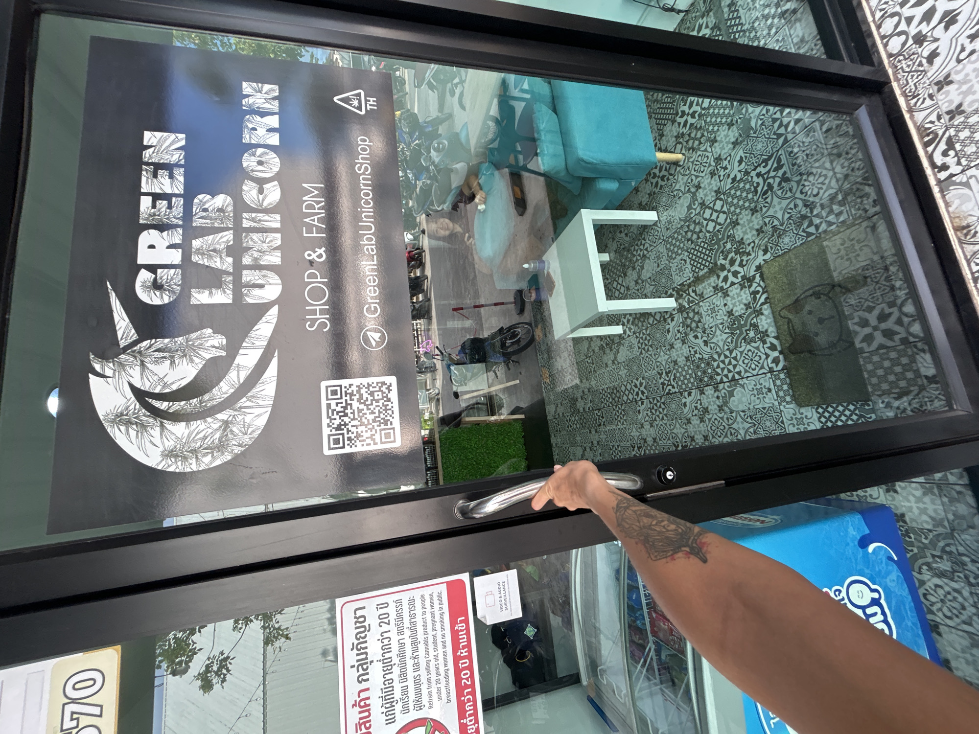 In Ratsada, Thailand, a person's tattooed arm reaches for a glass door handle. The door displays a prominent sign for "GREEN LAB UNICORN SHOP & FARM," featuring a unicorn illustration and the shop's name in large lettering. A QR code is also visible on the sign. Through the glass, the interior of the shop is visible, with a turquoise sofa and a white table. Outside, motorcycles are parked in a lot, and a woman is seated inside, partially visible through the reflection. A red and white sticker on the door states that cannabis products are prohibited for individuals under 20 years old, students, pregnant women, breastfeeding women, and those who are smoking in public.