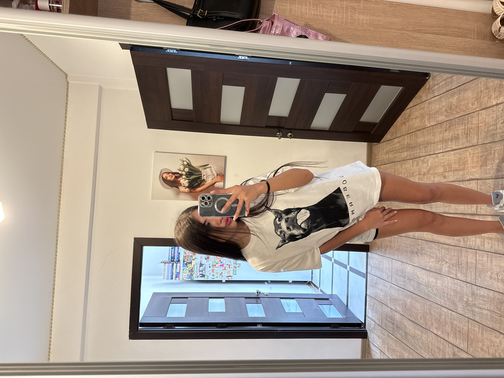 A young woman with long, dark hair is taking a mirror selfie in a hallway. She is wearing a white t-shirt with a black dog graphic and black shorts. She is holding a smartphone in her right hand. To her left, there is a dark wooden door with glass panels, open to reveal a brightly lit room with a checkered floor. To her right, another dark wooden door is closed. The walls are a neutral color, and there is a framed picture of a woman with flowers on the wall. A shelf unit with books and other items is visible on the far right. The floor of the hallway is made of light-colored wood planks. The scene appears to be an indoor residential setting in Chișinău, Moldova.