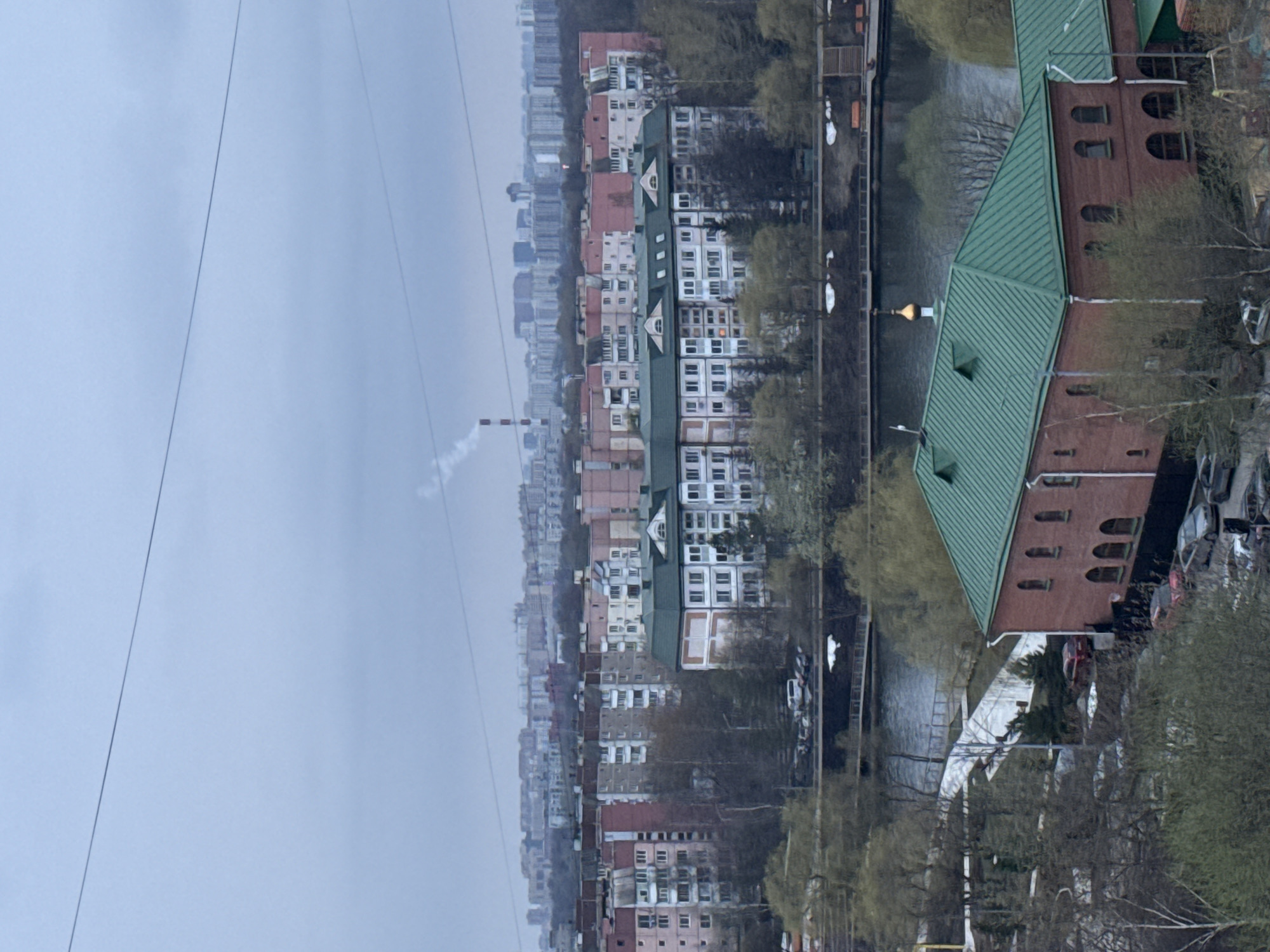 The image depicts a cityscape, likely Moscow, Russia, on an overcast day. In the background, a long row of apartment buildings of varying heights and colors extends into the distance. A smokestack emitting a plume of smoke is visible among the buildings. To the right, a river or canal runs parallel to the buildings. A red-brick building with a green, peaked roof is in the foreground along the river's edge. Sparse, bare trees line the riverbank. Cars are parked along the road next to the river. Overhead, electrical or telephone wires traverse the sky.