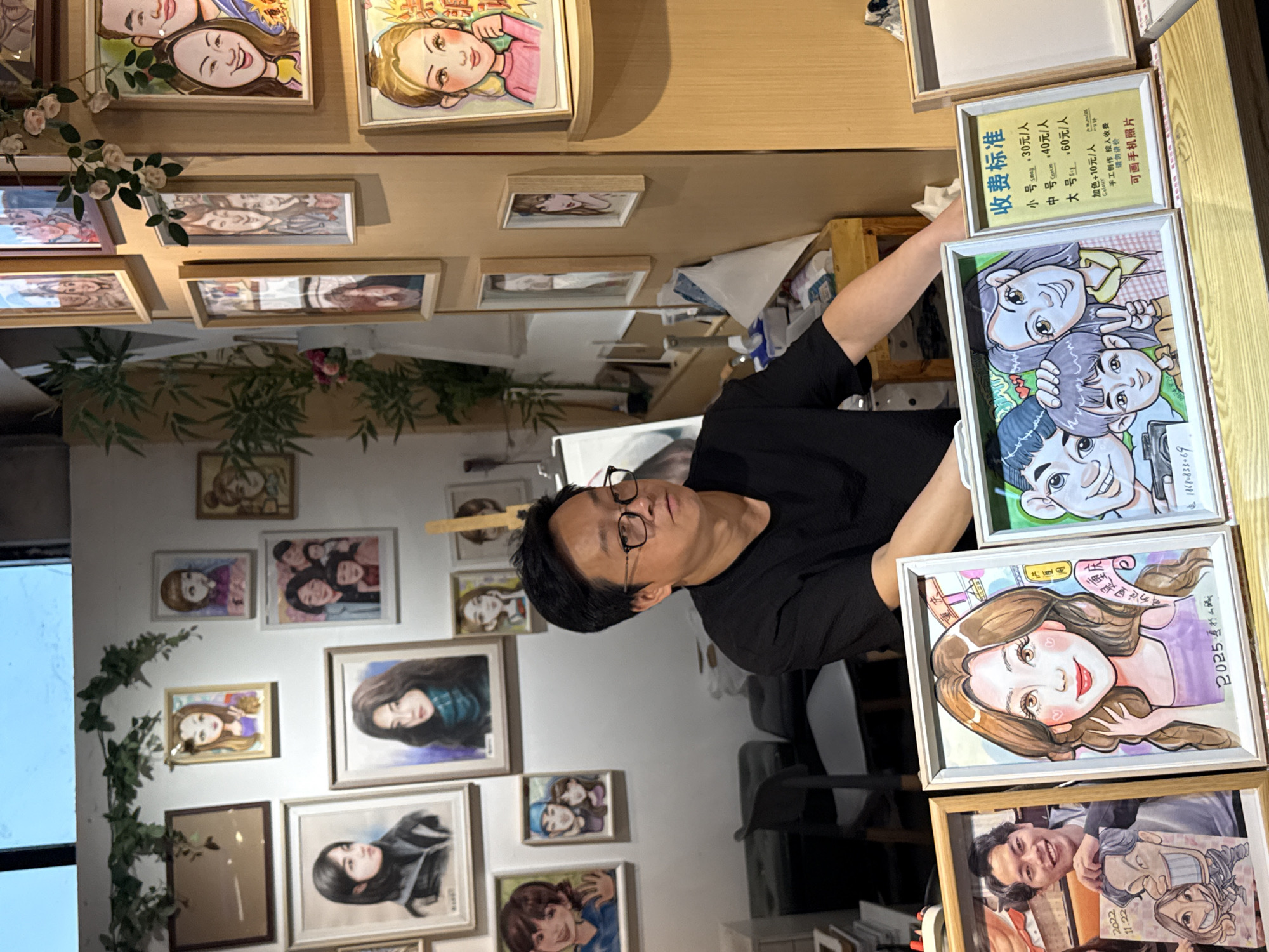 The image captures a scene in what appears to be an art studio or a space showcasing portrait art, likely located in the Yuzhong District of China. The wall is adorned with numerous framed portraits, varying in size and style, some of which are caricatures and others realistic paintings. An Asian man wearing glasses and a black t-shirt is standing and holding a stack of framed portraits. One of the frames displays what appears to be a pricing chart, written in Chinese, suggesting the cost for different sizes of commissioned portraits. The scene is somewhat cluttered, with the artistic displays taking center stage. The man's interaction with the portraits, combined with the presence of the pricing chart, hints at a business related to creating and selling portraits.