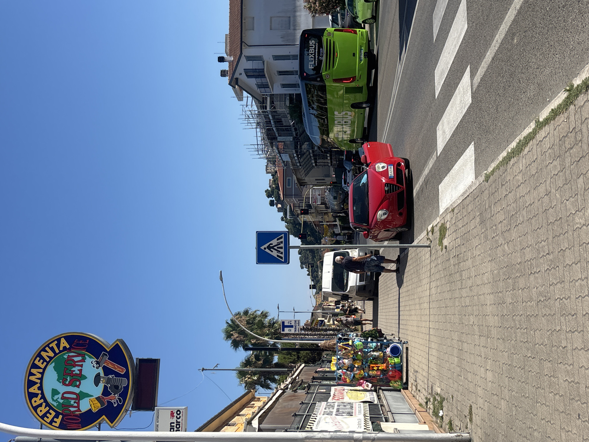 This is an eye-level, medium shot of a street scene in Cupra Marittima, Italy, on a sunny day. On the left side of the image, a sidewalk lined with palm trees and buildings is visible. A street vendor's cart, stocked with various colorful items, is positioned on the sidewalk. Beyond the cart, a white van and a red Alfa Romeo sports car are parked or moving. Across the street, a large green FlixBus coach is traveling in the right lane. The background shows buildings under construction, some with scaffolding, and more distant hillside homes. The sky is clear and blue.