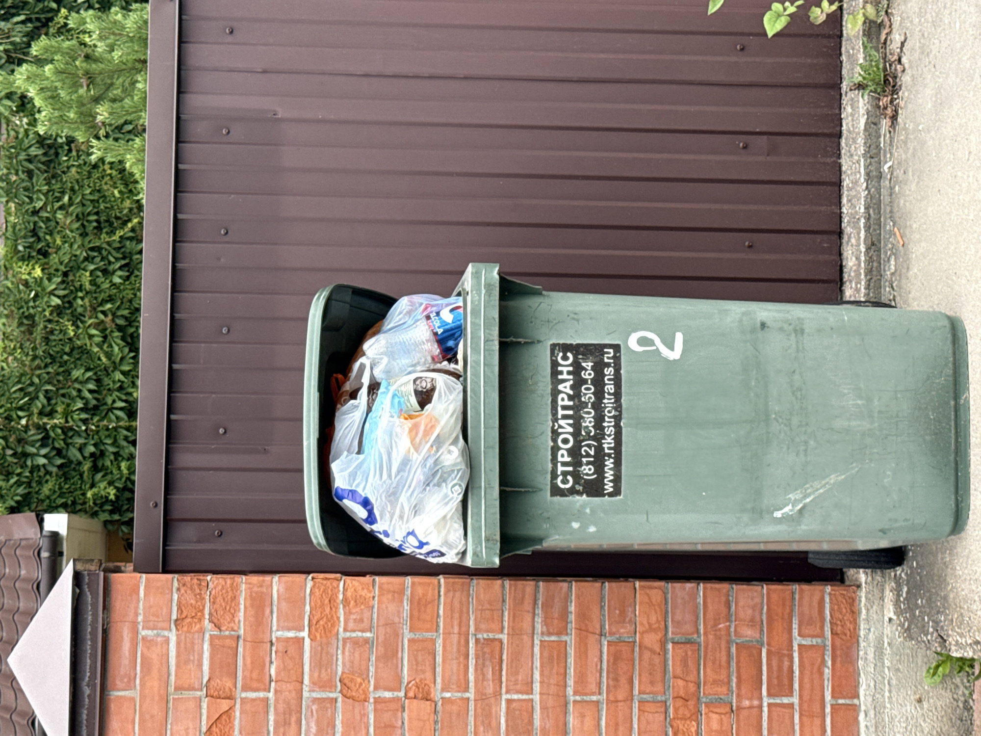 An outdoor medium shot shows a green, lidded garbage bin overflowing with plastic bags of refuse. The bin is situated in front of a brown, corrugated metal fence, with a brick column on the left. A large, green leafy plant is visible to the right of the bin. A white numeral "2" is painted on the front of the bin, above a black sticker that displays text in Cyrillic, a phone number, and a website address. The ground in front of the bin is concrete, with some small weeds growing. The overall setting appears to be residential or commercial.
