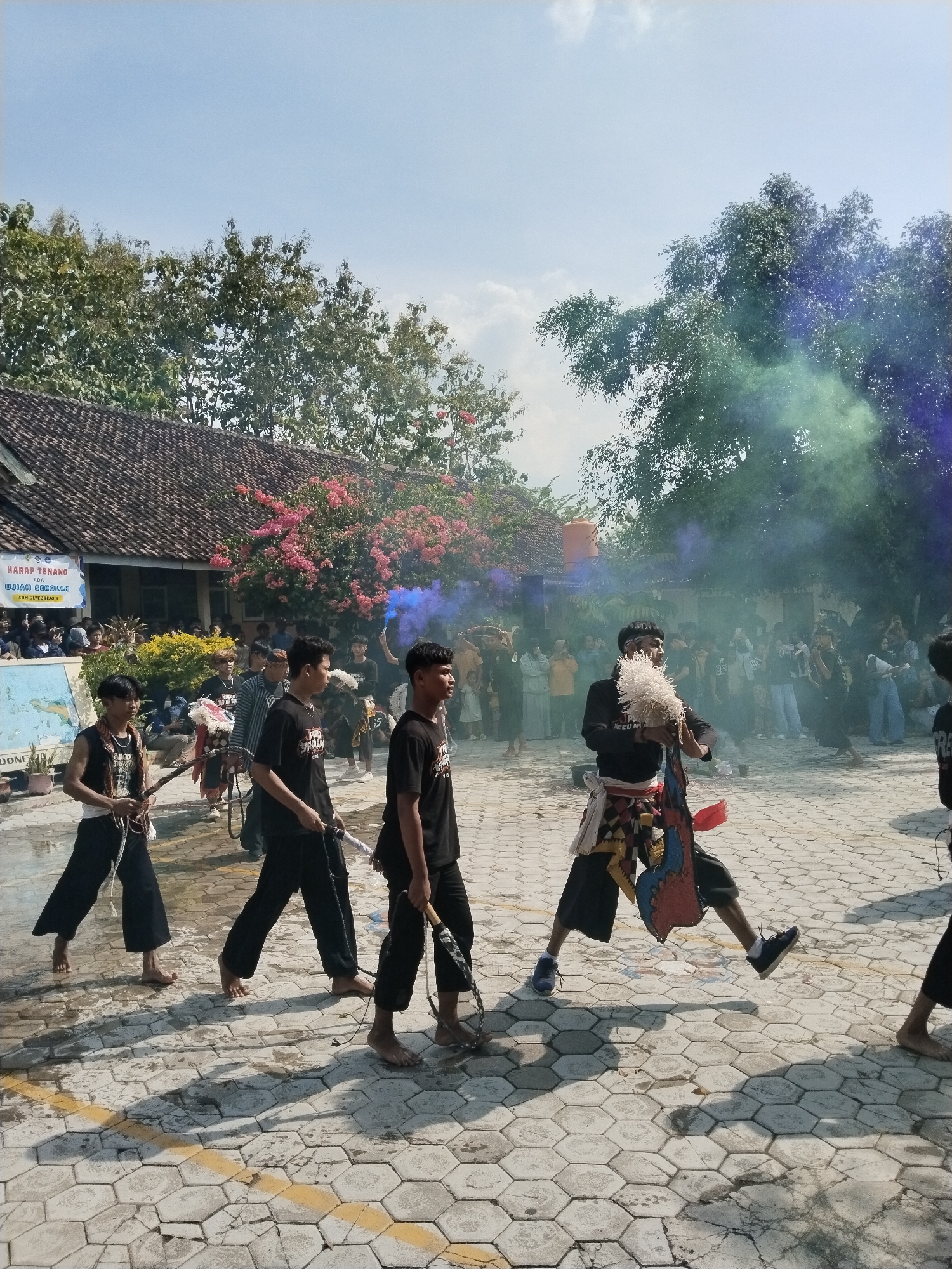 The image shows a cultural performance in Karangmalang, Indonesia. A group of young men are the focus, some barefoot, wearing black shirts and pants, and carrying whips or other implements. One performer is mid-leap, wearing shoes, an ornate waistcloth, and holding a feather-like object. They are performing on a paved area. In the background, a crowd is watching, and blue and green smoke is visible. A building with a tiled roof and bougainvillea flowers is present, along with a sign that reads "HARAP TENANG ADA UJIAN SEKOLAH." A map is partly visible on the sign. The sky is blue with some clouds. Trees are present in the background.