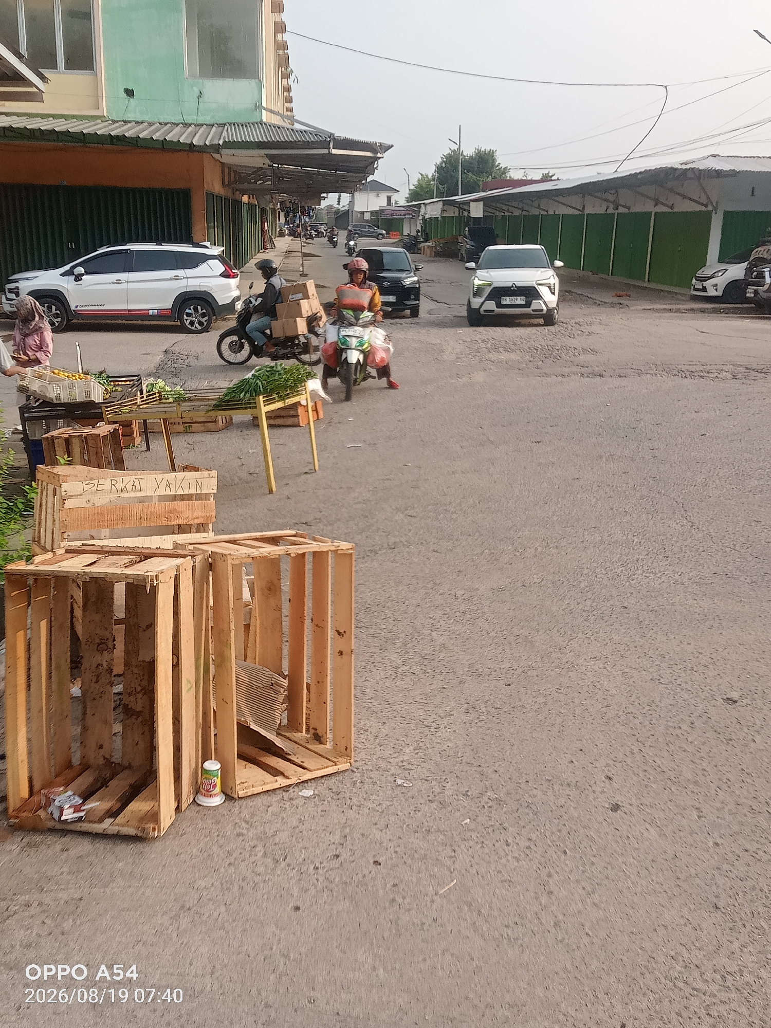 This is a wide-angle, eye-level shot of a street scene in Jambi, Indonesia, likely a market area. The foreground is dominated by several empty wooden crates, some stacked and others scattered. A white cup with a colorful label rests inside one of the crates. Further back, a person on a white motorcycle with a red seat is riding away from the camera, carrying boxes stacked on the rear. Next to them, another person on a motorcycle is also moving away. To the left, a white SUV is parked, and a person in a pink headscarf is standing next to a stall laden with oranges and green vegetables. In the background, a long building with green panels and a sloped roof extends down the street, with more motorcycles and vehicles visible. The street is paved and appears to be in moderate condition, with some cracks and unevenness. The sky is overcast, suggesting a hazy or cloudy day. The timestamp on the image reads "2026/08/19 07:40", indicating it was taken on August 19, 2026, at 7:40 AM. The text "OPPO A54" is also visible in the bottom left corner, likely indicating the device used to capture the image.