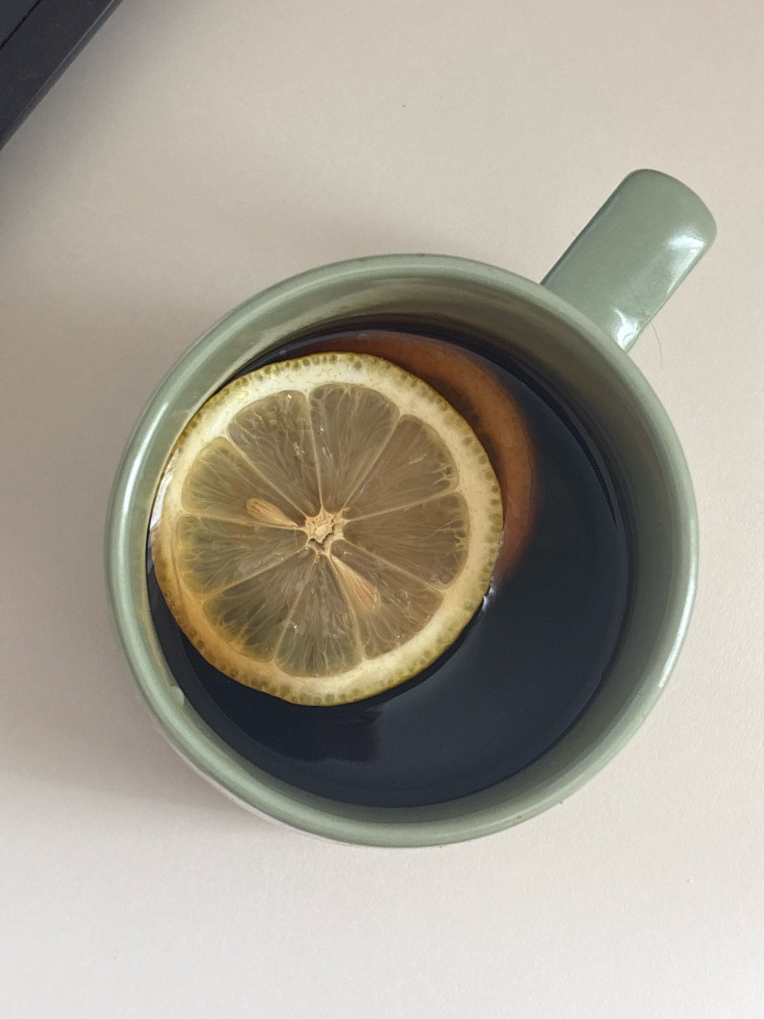 A close-up overhead shot shows a light green ceramic mug filled with dark liquid, likely tea or coffee. A slice of lemon is floating on the surface of the liquid, partially obscuring a slice of what appears to be orange. The mug is situated on a light-colored, matte surface. A portion of a dark object, possibly a laptop or a keyboard, is visible in the upper left corner of the frame, suggesting an indoor setting. The scene is clean and uncluttered, with no people present. There is no information in the image that could indicate a specific location in Bar, Montenegro.
