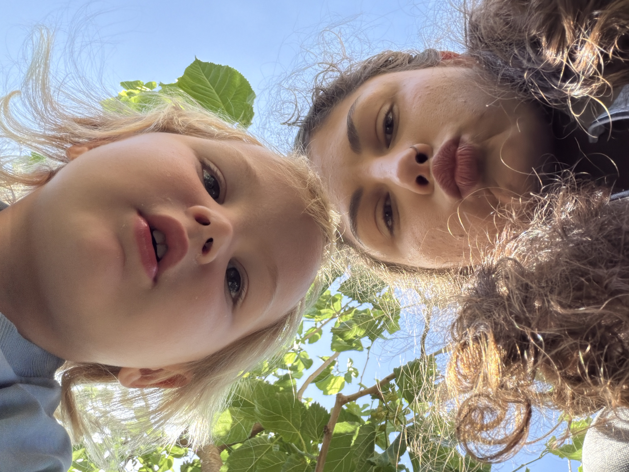 Two individuals, likely a young child with blonde hair and a young adult female with dark hair, are positioned closely together, looking downwards at the camera. Their faces occupy the majority of the frame. The child's mouth is slightly open, while the young adult female has her lips pursed. The background reveals green foliage and a glimpse of a blue sky, suggesting an outdoor setting. The angle of the shot indicates the photo was taken from below. No specific objects or actions are discernible, and the location in Kiryat Bialik, Israel, cannot be confirmed with certainty.