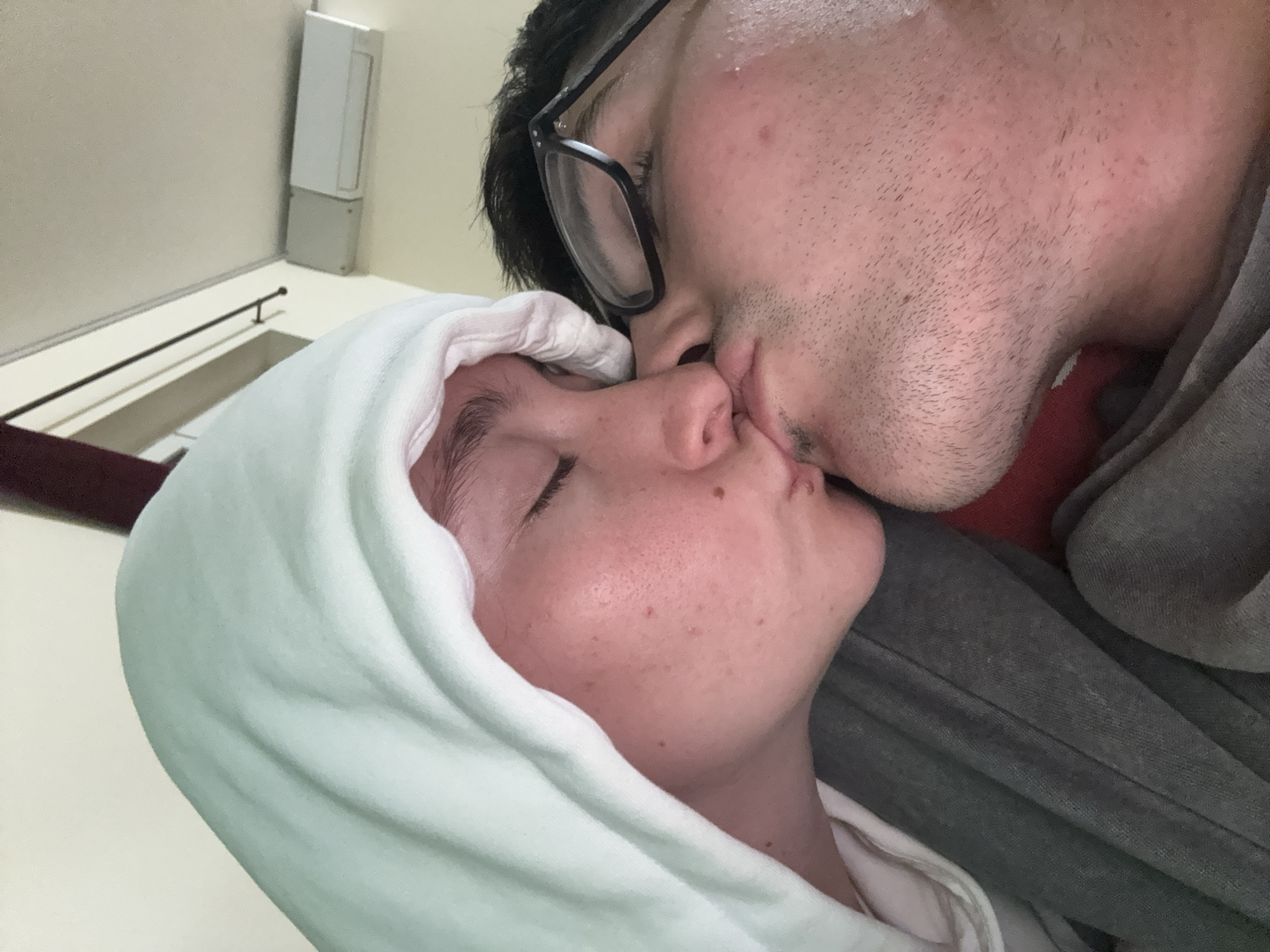 In Sofia, Bulgaria, a close-up, eye-level shot captures two individuals sharing a kiss. The person on the left, wearing a light blue hooded sweatshirt, has their eyes closed, and their face is angled slightly upwards. Their skin is fair with visible freckles and some redness. The person on the right, wearing glasses, has their eyes closed as well. Their skin appears slightly flushed, and stubble is visible on their chin and jawline. The background is blurred, but hints of a wall, ceiling, and an air conditioning unit are discernible, suggesting an indoor setting. The overall lighting is soft and warm, contributing to an intimate atmosphere.