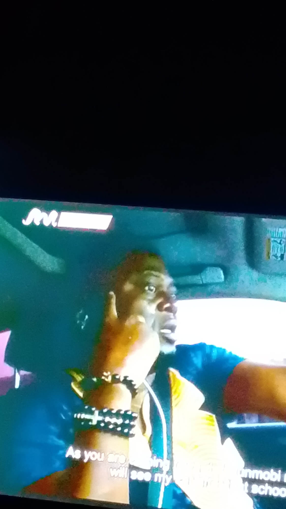 A man with dark skin and a short haircut is in the driver's seat of a vehicle, looking towards the right side of the frame with his mouth open as if speaking. He is wearing a blue shirt and a yellow garment over it, possibly a vest. On his left wrist, he wears two black beaded bracelets, one with a cross-shaped charm. Subtitles are visible at the bottom of the image. A logo or graphic with "M!" is present in the upper left corner of the frame.
