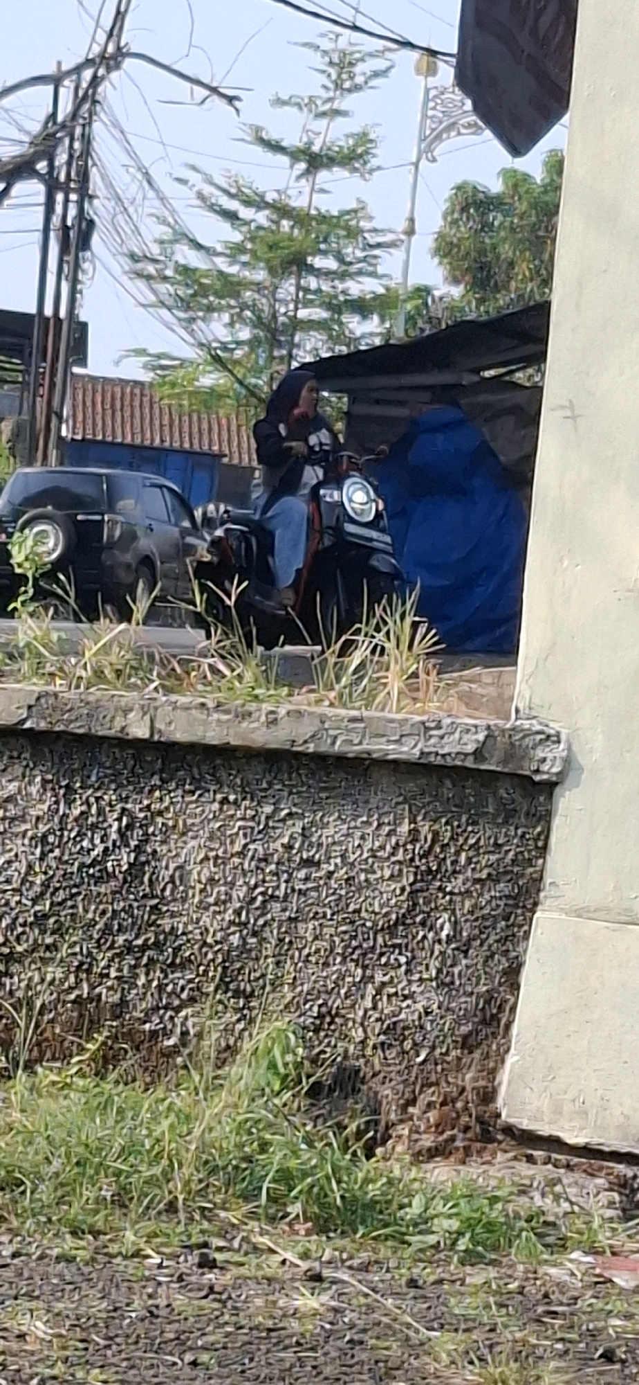 In Cilaku, Indonesia, a woman wearing a black hijab and a black jacket over a white shirt, with blue jeans, is riding a black motorcycle. She is positioned on the right side of the road, facing forward. Behind her motorcycle, a black SUV is visible. The background features a street light, electrical wires, trees, and a building with a large blue tarp. The scene is captured from a slightly elevated perspective, looking down towards the road. The foreground shows overgrown green grass and a textured stone wall.