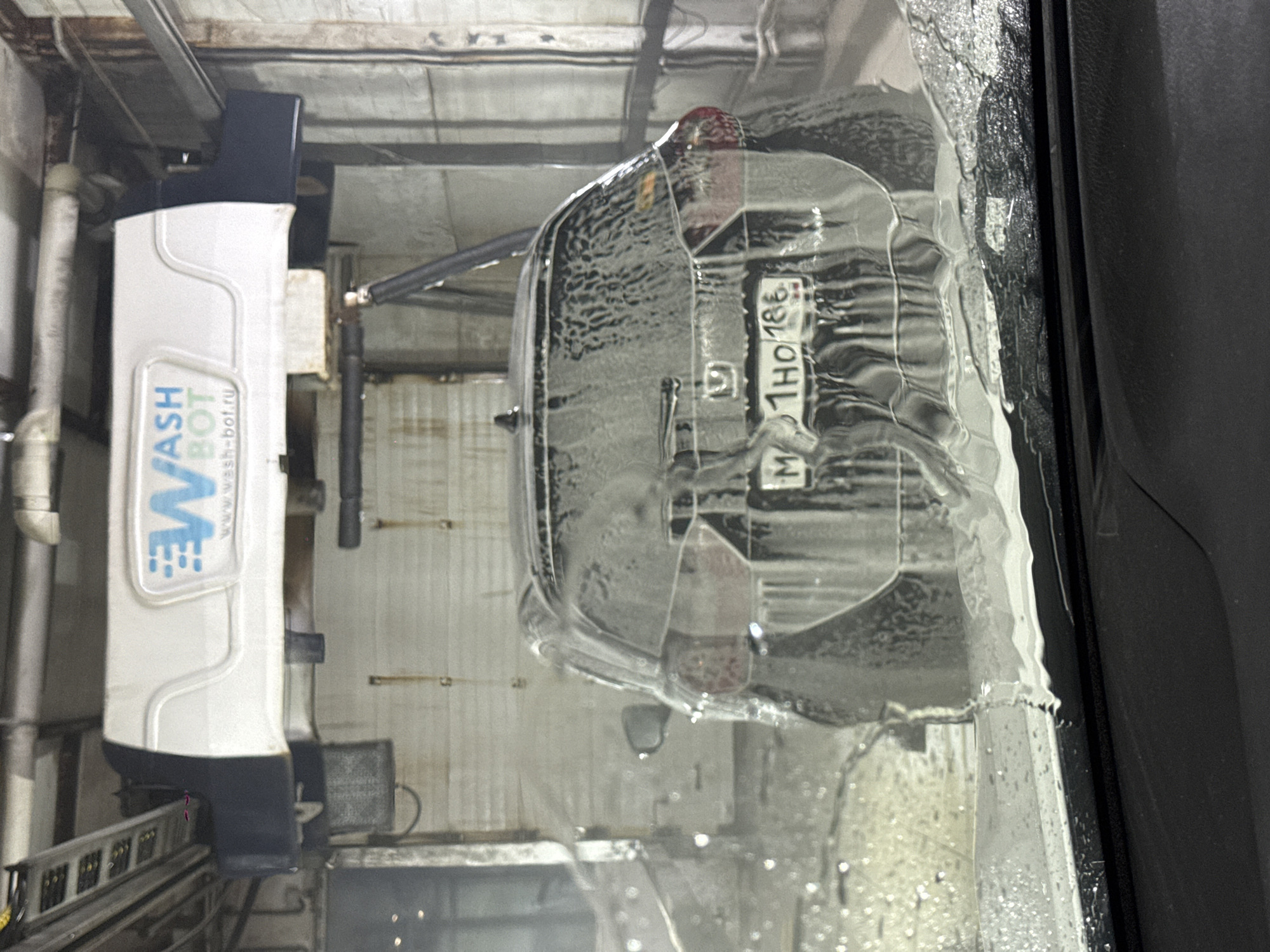 The image shows the rear of a car undergoing a wash in what appears to be an automated car wash. The car's rear window, partially obscured by water droplets, reveals the license plate "M 1HO 186". To the left, part of the car wash machinery is visible, labelled "WASH BOT" with the website "www.wash-bot.ru". The overall environment suggests an indoor car wash facility, possibly located in Nizhnevartovsk, Russia, based on the license plate format.