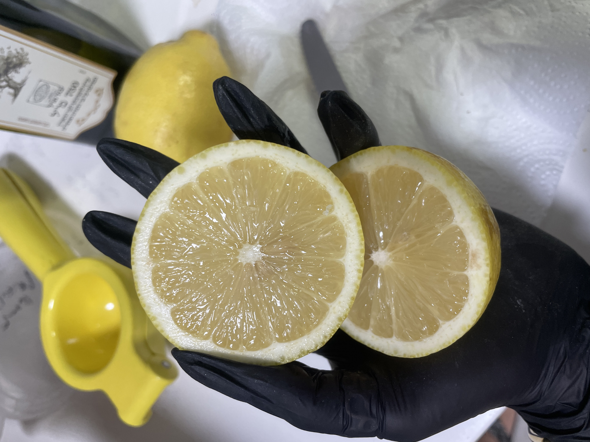 A person wearing a black glove holds two halves of a lemon over a white paper towel. In the background, there is a yellow lemon squeezer, another whole lemon, and a bottle of olive oil with a label in Hebrew. The scene is indoors, with a neutral background. The lighting is bright and direct, creating shadows from the objects. The image appears to be a close-up shot, focusing on the lemons and the hand. The location in Raanana, Israel, is not evident from the visual information in the image itself.