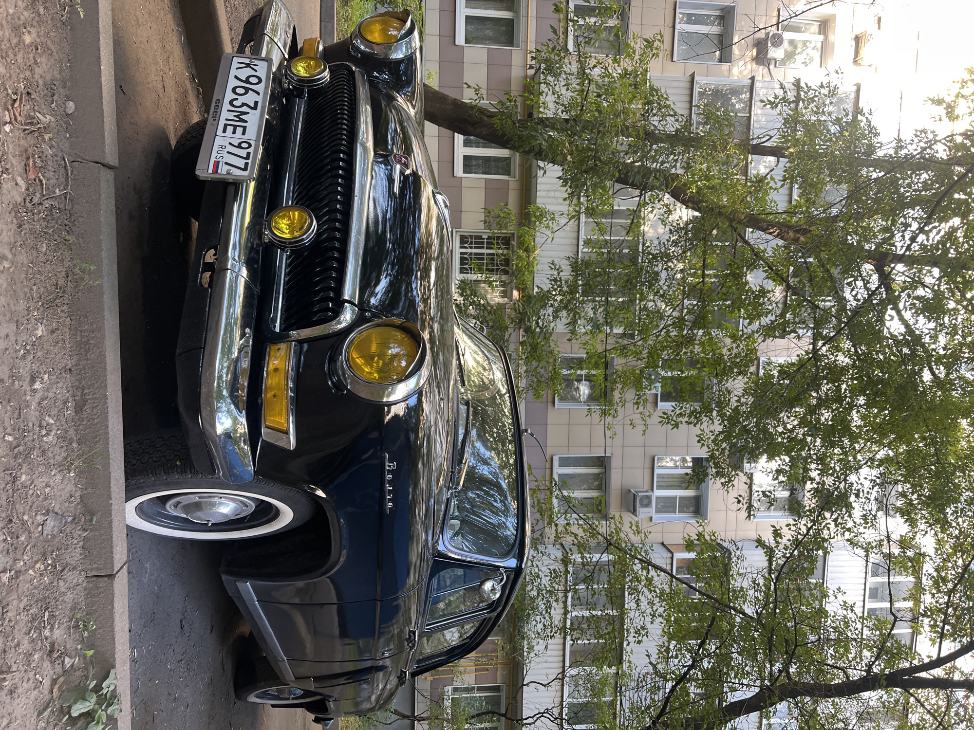 In Moscow, Russia, a vintage black Volga GAZ-21 car, manufactured between 1956 and 1970, is parked on a paved surface next to a sidewalk. The car features distinctive yellow headlights, chrome accents, and whitewall tires. In the background, a multi-story apartment building with numerous windows and balconies is visible, partially obscured by the foliage of a mature tree. The sunlight filters through the leaves, casting dappled shadows on the car and the building. The license plate of the car reads "K963ME 977" with "CCCP" and "RUS" markings. The environment appears to be a residential area in Moscow.