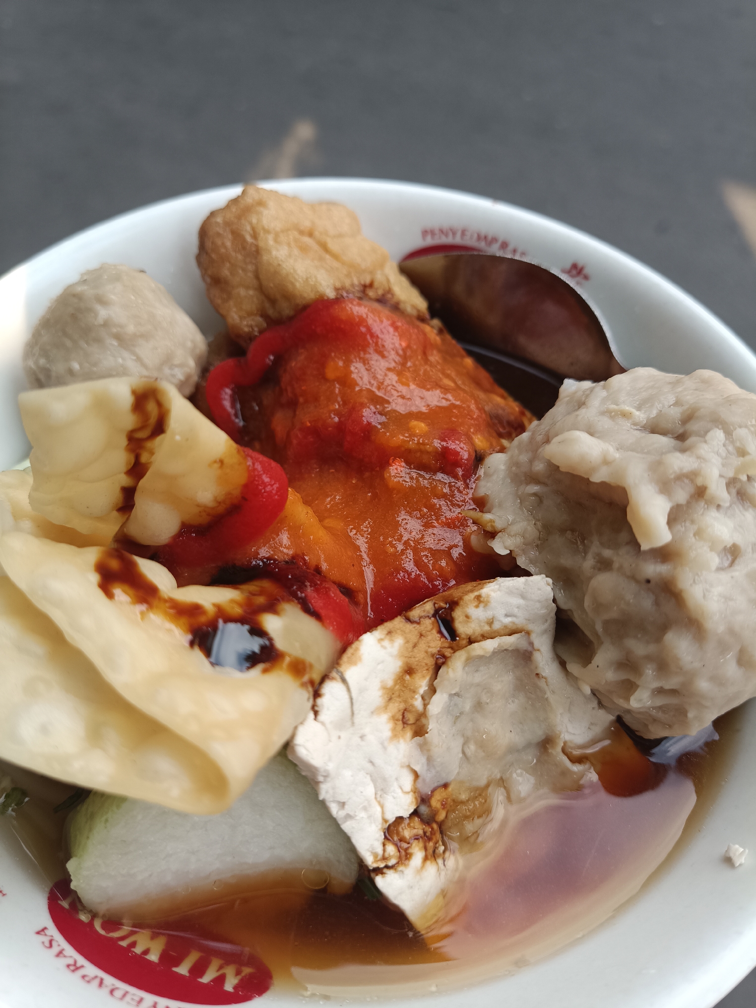 A close-up, high-angle shot captures a bowl of Indonesian meatballs, known as bakso. The bowl is white with red lettering that appears to say "NYEDAP RASA" and "MI WOK". Inside the bowl, several large, pale meatballs are visible, along with pieces of fried tofu, a few wonton wrappers drizzled with dark sauce, and a mound of red chili sauce. A spoon is partially submerged in the broth. The background is out of focus, showing a gray, textured surface, possibly a table or pavement. The lighting suggests it is daytime, with some light reflecting off the broth and spoon. This dish is commonly found in food stalls and restaurants across Indonesia, including the city of Jember, East Java, where it is a popular street food.