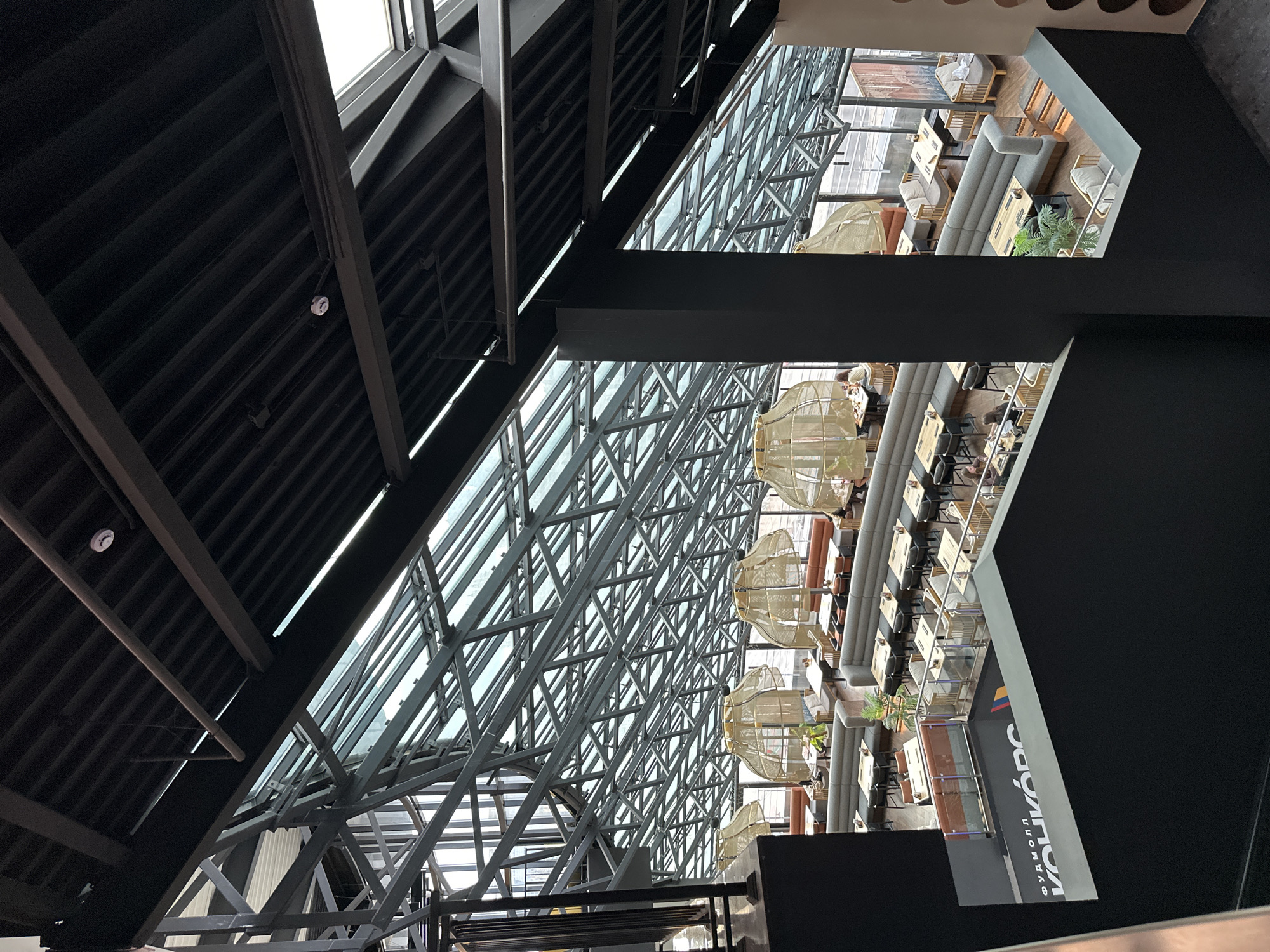 This image shows an interior view of a modern, multi-level restaurant or cafe with a distinctive glass and metal ceiling structure. The space is filled with natural light filtering through the expansive roof. The layout features rows of tables and seating areas, including booth-style seating along curved walls and individual tables with chairs. Large, woven pendant lamps hang from the ceiling, adding a warm, decorative element. Several patrons are visible, seated at tables, engaged in dining or conversation. The overall aesthetic is contemporary and airy, with a focus on architectural design. The sign at the bottom left of the image displays "ФУДМОЛЛ КОНКОРД" which translates to "Food Mall Concord," identifying the location as the Food Mall Concord in Minsk, Belarus.