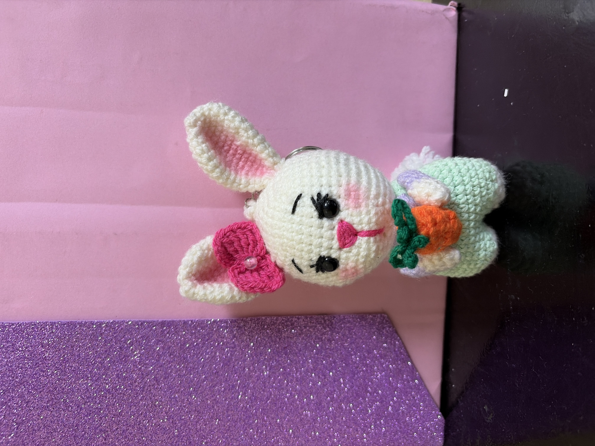 A close-up shot features a crocheted rabbit keychain lying on a textured surface. The rabbit is primarily white with pink accents inside its ears and on its cheeks. It has a pink bow attached to one ear. The rabbit has black eyes and a small pink nose. Behind the rabbit's head is a small, crocheted green object with an orange carrot attached. A black fuzzy ball is attached to the back of the green object. The background consists of layers of different materials including a pink layer, a purple glitter layer, and a dark purple shiny layer. No information on the location is available.