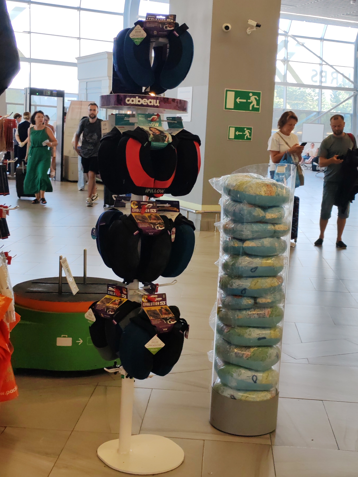 In a well-lit airport terminal, a retail display showcases travel pillows. The display features a variety of neck pillows, some in black and red, others in dark blue and green, with packaging indicating the "Evolution S3" and "iPILLOW" brands. Nearby, a cylindrical rack is filled with stacked travel pillows featuring a world map design.
Several people are visible in the background. A woman in a green dress is walking, pulling a suitcase. A man with a beard, wearing a dark t-shirt and shorts, is also walking. Further back, a woman in a white t-shirt looks at her phone, and a man in a grey t-shirt stands nearby, holding a bag.
The setting includes an airport environment with large windows, a metal detector, and directional signage indicating exits. The flooring is tiled. The time of day appears to be daytime, given the natural light. The overall atmosphere suggests a busy travel hub. Visible text includes brand names like "cabeau" and product names like "EVOLUTION S3" and "iPILLOW," as well as directional arrows and icons related to exits and luggage.