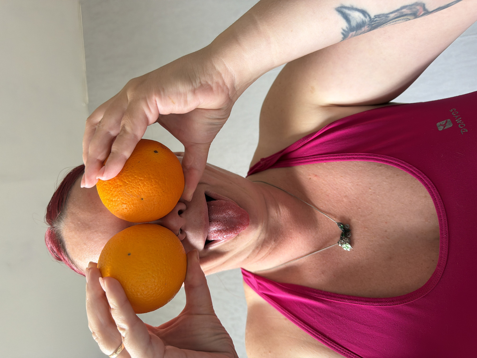 A person is holding two oranges in front of their face, covering their eyes, with their tongue sticking out. The person has reddish-purple hair and is wearing a pink tank top and a silver necklace. There is a tattoo on their left arm. The background is a plain, off-white wall. It's impossible to determine the location of this image with only this information.