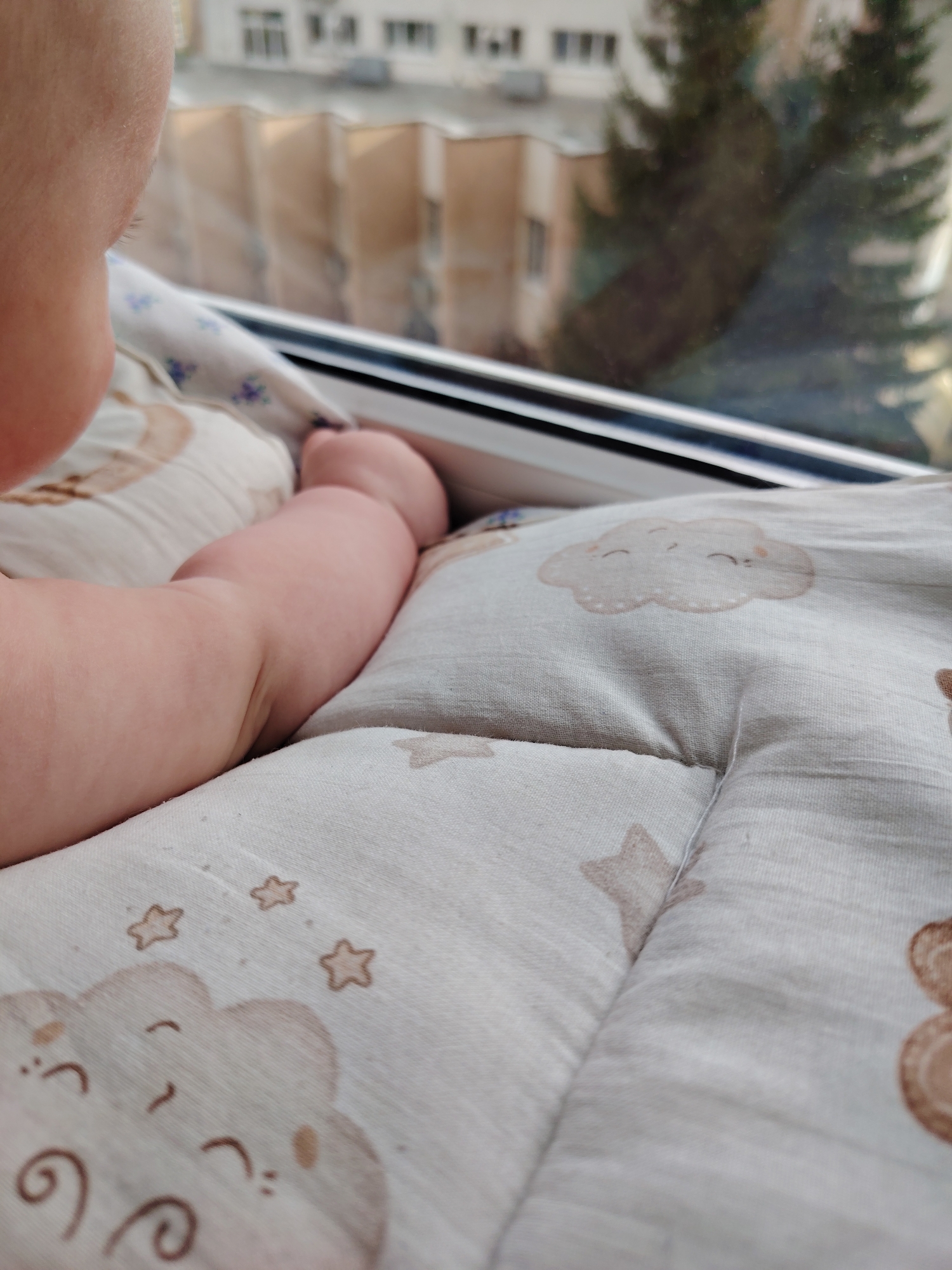 An overhead, medium shot shows a baby's arm resting on a quilted blanket with a cloud and star pattern. The blanket is positioned near a window, with a blurred cityscape and evergreen tree visible through the glass. The baby's head and shoulder are partially visible in the upper left corner of the frame. The scene appears to be indoors.