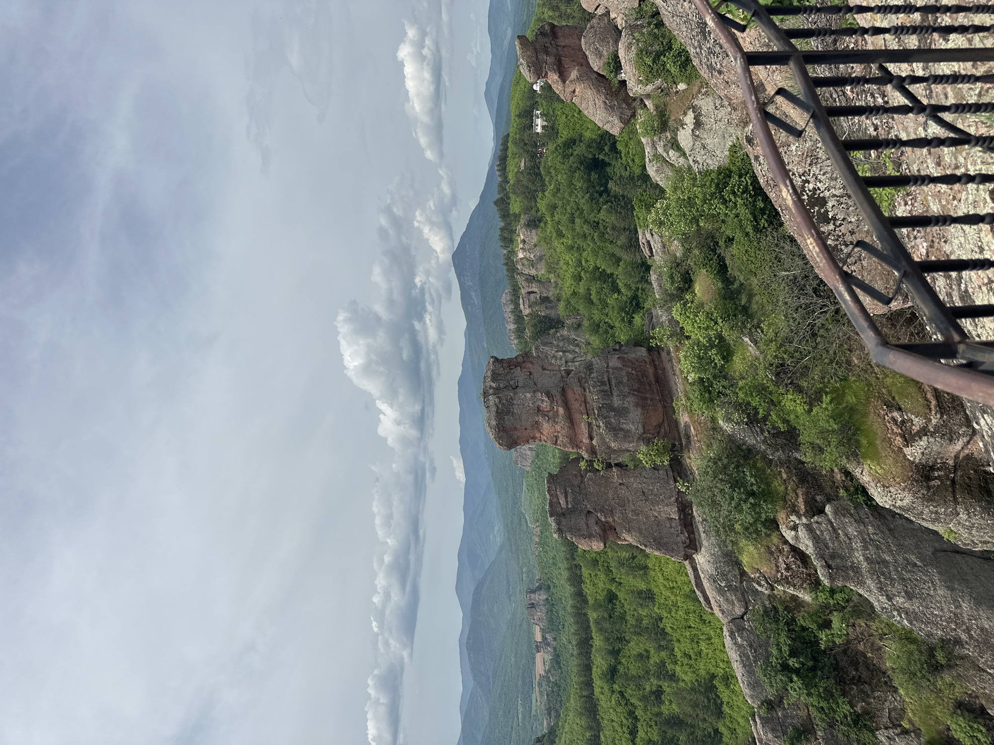 This image captures a view of the Belogradchik Rocks in Bulgaria. The scene includes large, reddish-brown rock formations covered with some greenery and trees. There's a green forested area spanning the base of the rocks and extending into the distance, where mountain ranges can be seen under a cloudy sky. A metal railing is visible in the foreground.