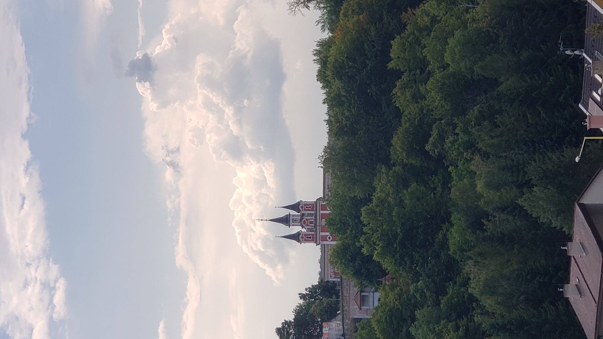 The image shows a view from a slightly elevated position looking towards a red and white building with three prominent spires, resembling a church or monastery, situated atop a hill covered in dense green trees. The sky above is partly cloudy with large, puffy white clouds against a pale blue backdrop. In the foreground, the roofs of several houses with chimneys are visible. This building is the Catholic Church of the Sacred Heart of Jesus in Prešov, Slovakia, also known as the Greyfriars' Church.