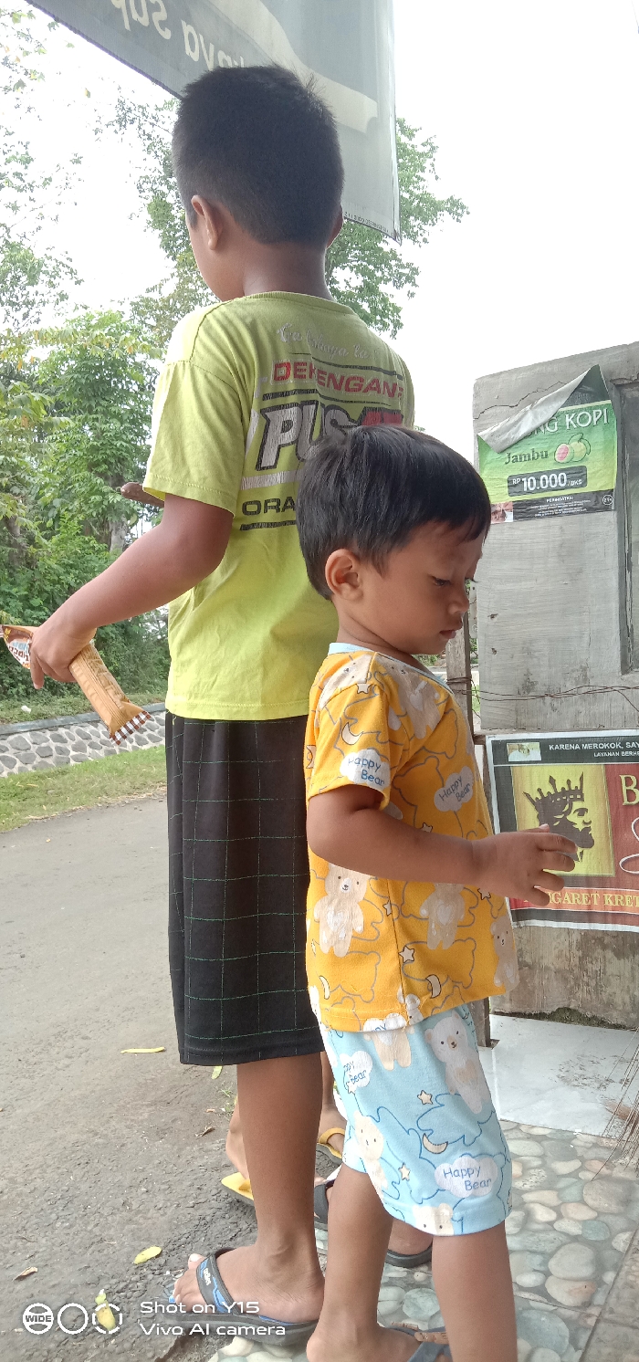 Two young children are standing next to each other. The older child is wearing a lime green t-shirt with black shorts, holding a wrapped snack. The younger child is wearing a yellow t-shirt with a teddy bear pattern and light blue shorts, also with a teddy bear pattern. Behind them is a weathered wall with posters and advertisements. The bottom left corner displays camera settings: "Shot on Y15 Vivo AI camera." The scene appears to be outdoors, possibly near a roadside shop or stall.
