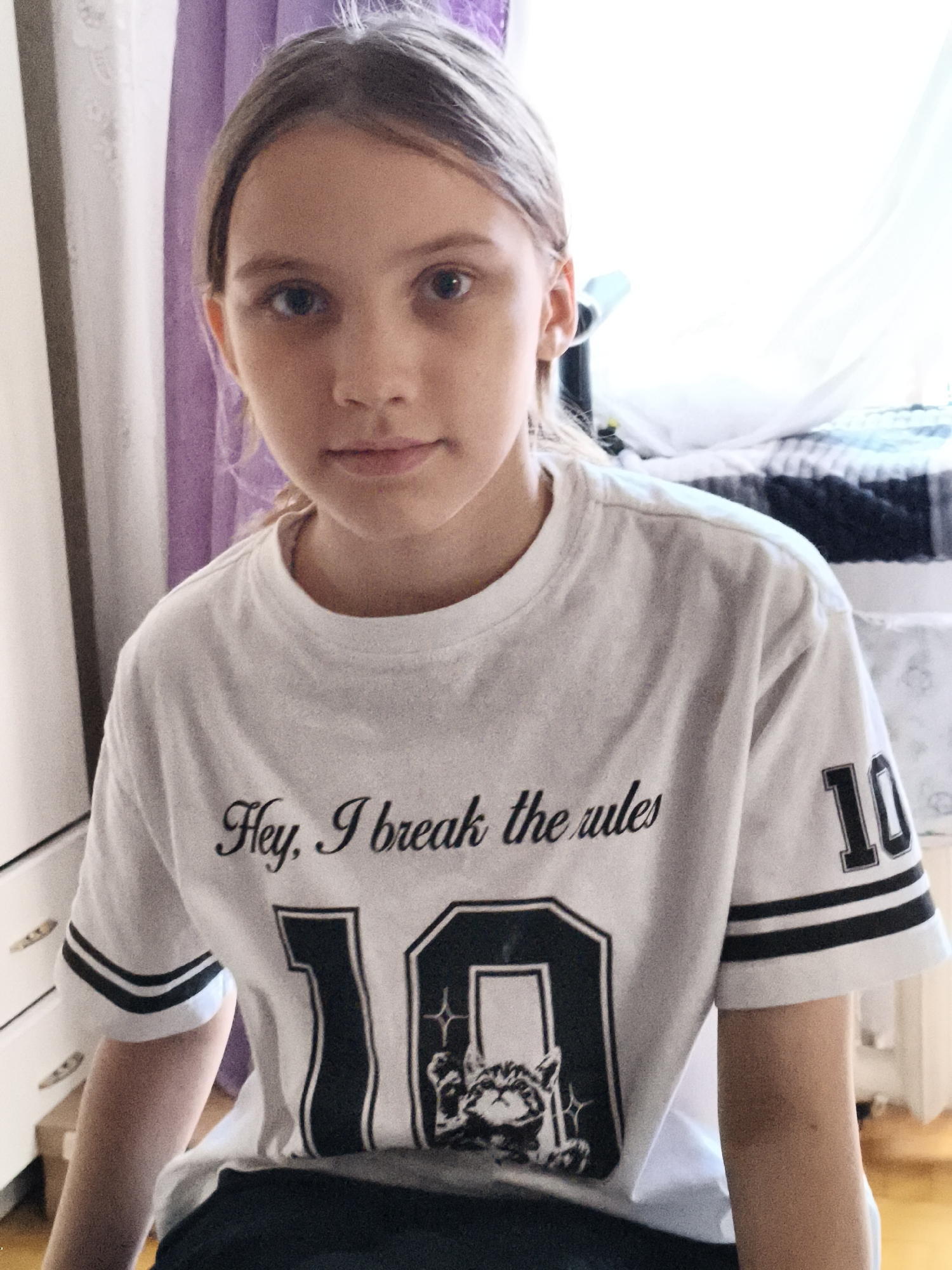 A young girl with light brown hair is posing for a portrait, looking directly at the camera. She is wearing a white t-shirt with a graphic design and text. The text on her shirt reads "Hey, I break the rules". The graphic features a large number "10" with a cat illustration inside. The background suggests an indoor setting with a dresser or cabinet to the left, purple curtains, and a bed with a black and white striped duvet to the right. The lighting indicates it is daytime, possibly late morning or early afternoon, as natural light streams in from a window, creating a bright atmosphere. The girl's expression is neutral.