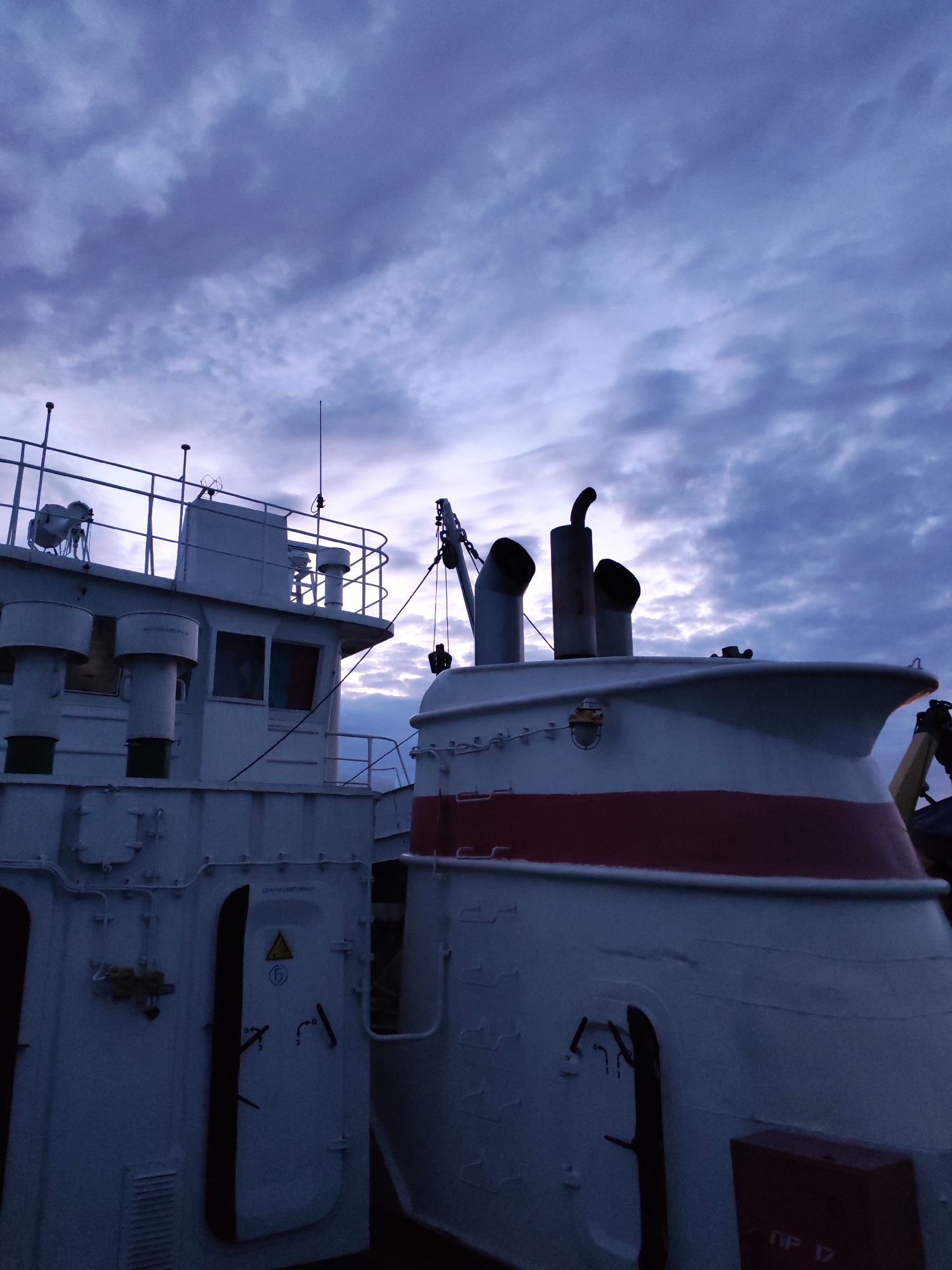 The image presents a dramatic maritime scene featuring two large vessels against a cloudy, twilight sky, likely at dusk or dawn. The overall lighting is dim, casting a cool blue and purple tone across the scene.
On the left, the white superstructure of a ship dominates a significant portion of the frame. It features a bridge or command deck with dark rectangular windows and a metal railing along its top, where an antenna and some equipment resembling life rafts are visible. Several cylindrical vents or small stacks rise from this deck. A prominent white door on its side displays a yellow warning triangle with an exclamation mark and a circular symbol. Above this door, Cyrillic text reads "АВТОНОМНЫЙ ПУСК" (Autonomous Start).
To the right, the bow or stern section of another ship is partially visible, also painted white but distinguished by a bold red stripe running horizontally along its hull. This vessel features two large, dark funnels or exhaust stacks and a small crane or derrick arm. A single, round light fixture is mounted on its side. In the lower right corner, a red box is marked with the white Cyrillic text "ПР 17" (likely an abbreviation for Project 17 or similar).
The sky above is filled with dense, textured clouds, ranging in color from dark blue to purple and lighter grey, with a brighter area visible towards the horizon. The absence of direct sunlight and the color palette strongly suggest either sunrise or sunset. There are no people visible, and no specific activity is taking place, contributing to a serene and still atmosphere. Based on the visible Cyrillic text, the location is in Russia, but the exact setting remains unknown.