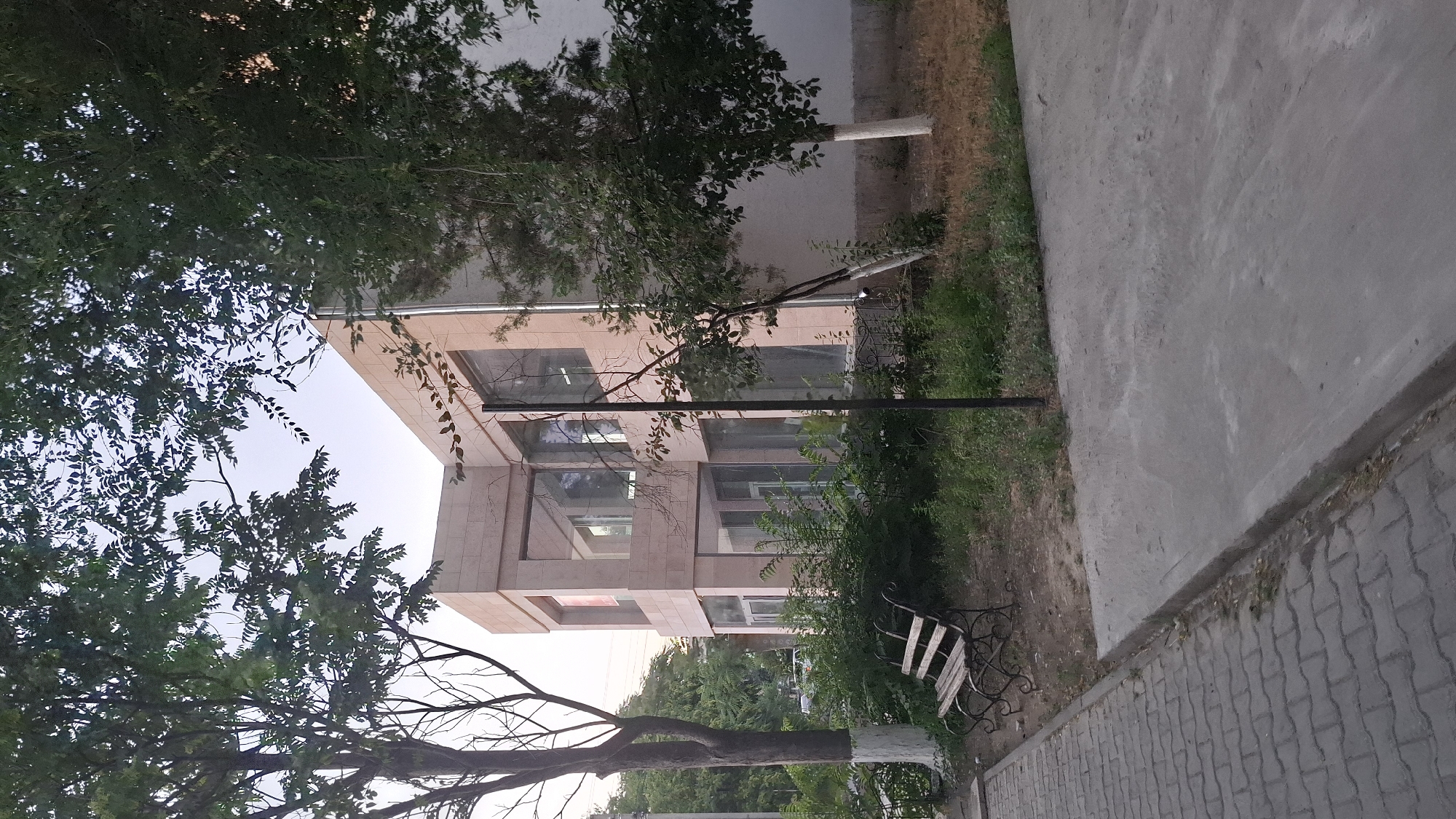 A modern, multi-story building with a light-colored facade and large windows is partially obscured by trees. In front of the building, a section of a paved walkway and a patch of green grass are visible. A metal bench with wooden slats sits on the grass area. The image is taken from a low angle, looking up towards the building and the sky, which is a pale blue. The trees are lush and green, with some branches extending across the frame. The lighting suggests it is daytime, possibly late afternoon, with the sun casting dappled shadows. The scene appears to be an outdoor public space, perhaps a park or a courtyard adjacent to the building. There is no visible text in the image.