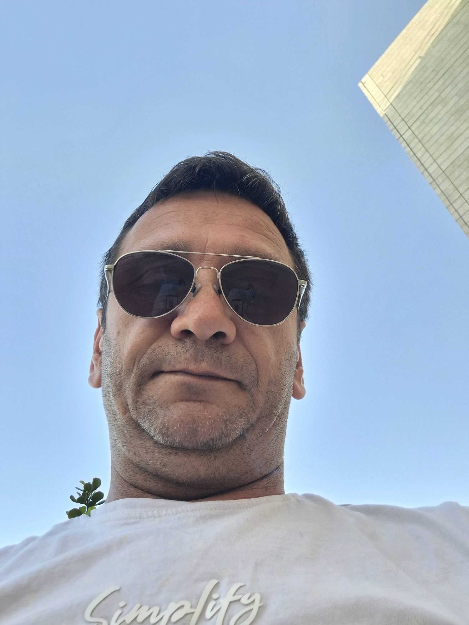 A low-angle, close-up, selfie-style photograph shows a man from the chest up, with the sky as the background. The man is wearing sunglasses and a white t-shirt. A small green plant is visible on his left shoulder, near his neck. The man's hair is dark and short, and he has facial stubble. A portion of a modern, light-colored building with grid-like markings is visible in the upper right corner of the image, against the blue sky. The context of Kiryat Gat, Israel, is absent from the visual information in the image.
