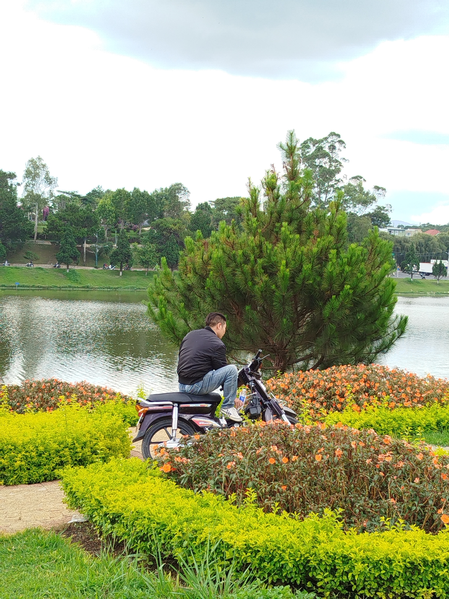 A man sits on a motorcycle by a lake in Da Lat, Vietnam. He is seen from the back, wearing a black jacket and jeans. The motorcycle is parked amidst a garden bed of green bushes and orange flowers. Behind him is a lake with trees on the opposite shore. The sky is overcast.