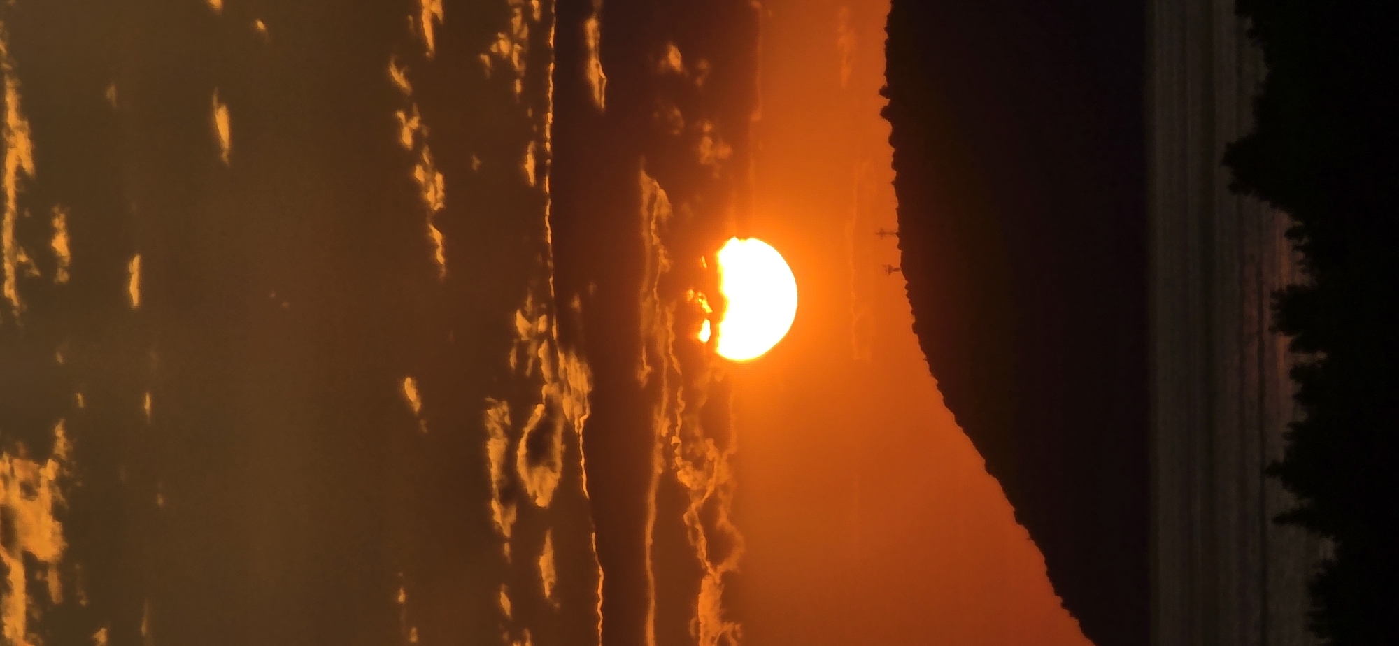 The image is an orange-toned landscape of a sunset in Bar, Montenegro. A bright sun is partially obscured by a dark, sloped silhouette, possibly a hill or mountain. The horizon line is visible, with a dark strip of water on the far right. The sky is filled with orange light, and there are some cloud formations on the left side. In the background, the silhouetted mountains are sparsely populated by the electric poles and lights.