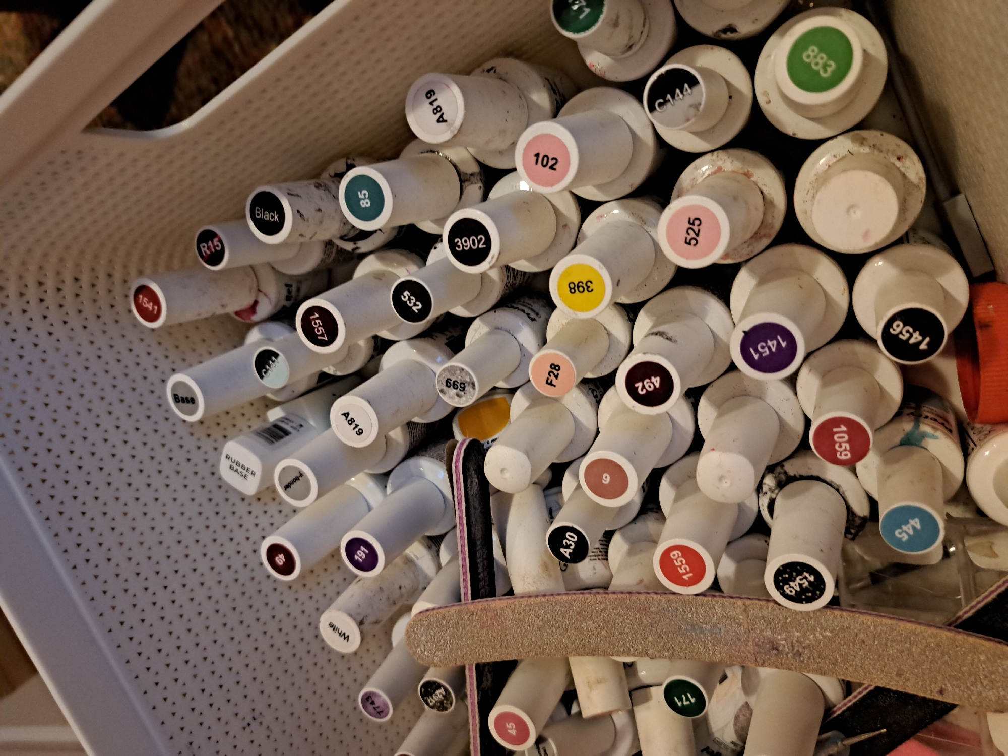 The image shows a collection of nail polish bottles in a white plastic basket. The bottles are various colors and appear to be gel nail polishes, judging by the labels. Some bottles have colored labels with numbers such as "102", "525", "966", "883" and "1451". Others feature labels with numbers such as "1549", "1559", "1059" and "445" or letters and numbers combinations like "A30". A nail file is among the bottles. There is no information provided that can confirm the location to be in Negovan, Bulgaria.
