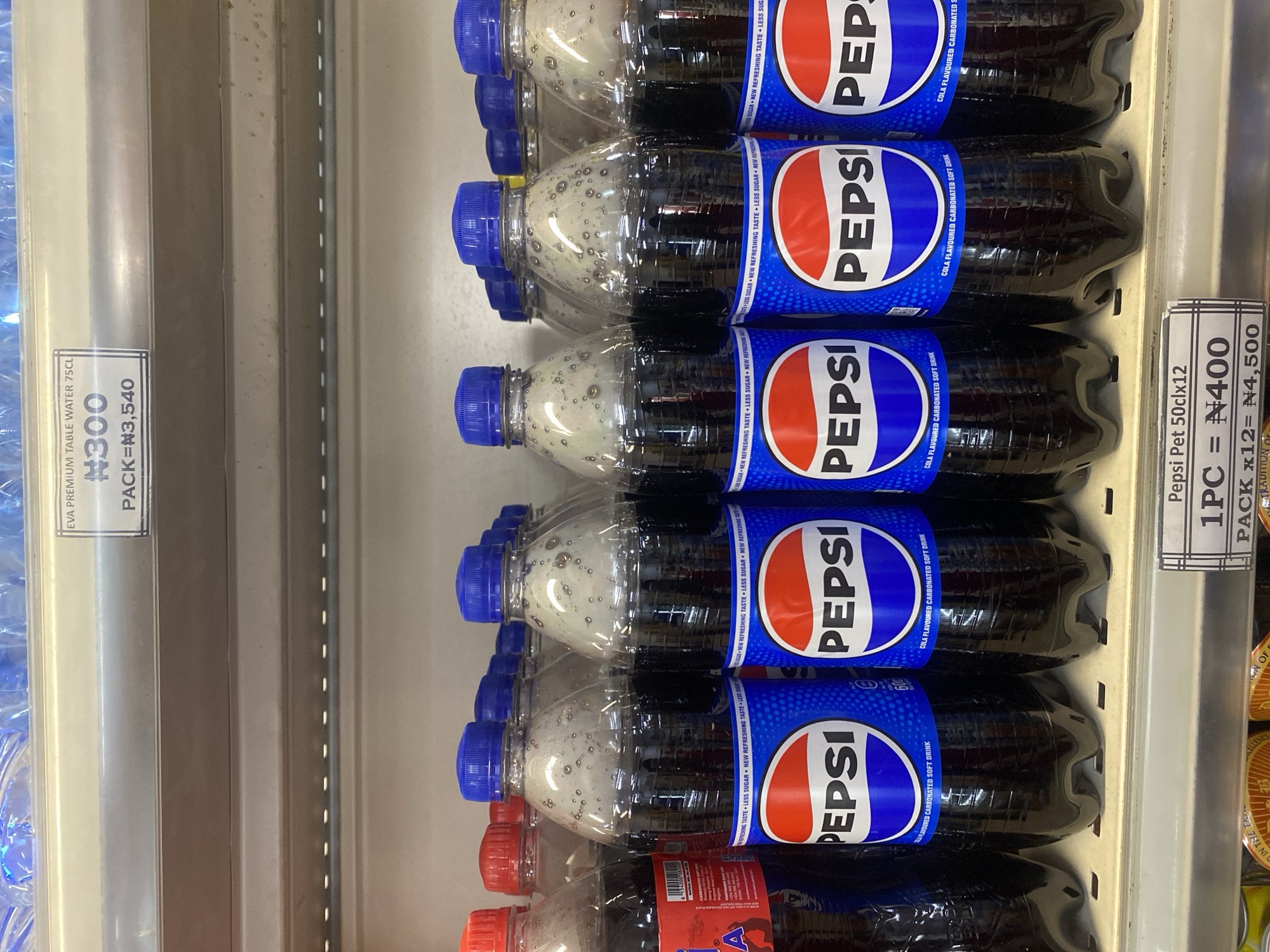 In Ikorodu, Nigeria, a retail shelf displays a selection of beverages. In the foreground, multiple plastic bottles of Pepsi, each with a blue cap and the iconic red and blue Pepsi logo, are lined up. Some bottles have visible bubbles inside. To the left, red-capped bottles, possibly also Pepsi, are partially visible. In the background, clear plastic bottles of water are stacked on a higher shelf. A white price tag above the water bottles indicates "EVA PREMIUM TABLE WATER 75CL" and a price of "#300 PACK = #3,540". Another price tag in the foreground reads "Pepsi Pet 50clx12 1PC = #400 PACK x12 = #4,500". The shelving unit appears to be made of metal, and the overall lighting suggests an indoor store environment.