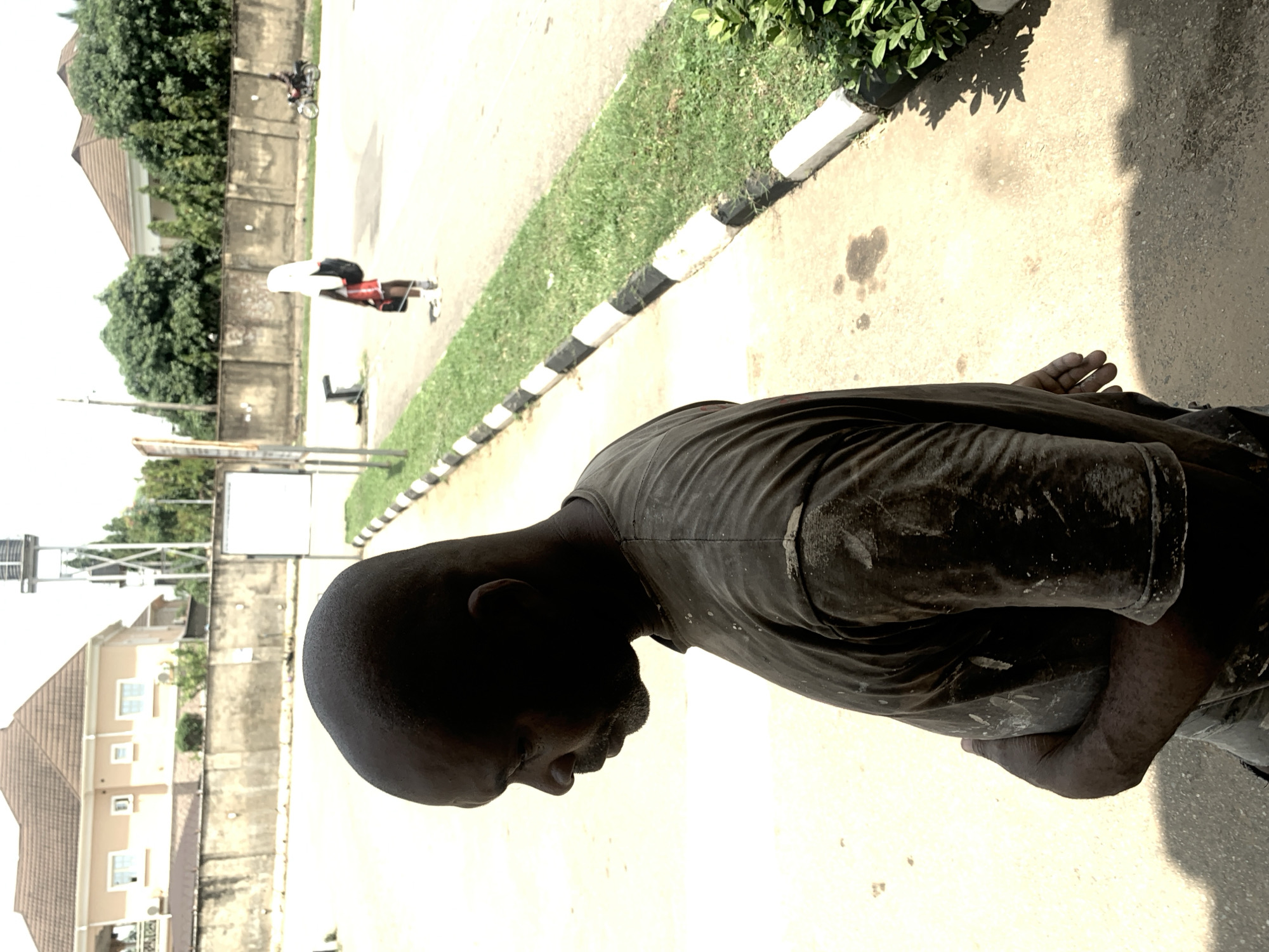 This media file, taken in Abuja, Nigeria, depicts a man in his 30s or 40s, with dark skin and a shaved head. He is wearing a dark t-shirt and dark pants, both appearing to be soiled with white specks, possibly paint or dust, suggesting he might be engaged in manual labor or construction. The man is shown from the chest up, with his head bowed and his face turned away from the camera, conveying a sense of weariness or introspection. His right hand is visible, resting on his lap, with his fingers slightly splayed.
The setting appears to be outdoors, with a paved walkway in the foreground, marked by a black and white striped curb. Behind the curb, a strip of green grass is visible. In the background, there is a concrete wall and beyond that, residential buildings with tiled roofs, characteristic of the area. Trees and foliage are also present, suggesting a somewhat suburban or residential environment. A tall water tower can be seen in the far background.
The lighting suggests it is daytime, possibly mid-morning or afternoon, with shadows cast on the ground. The overall atmosphere is neutral, with no strong indicators of a specific event or emotion beyond the man's contemplative posture. There is no visible text in the image.