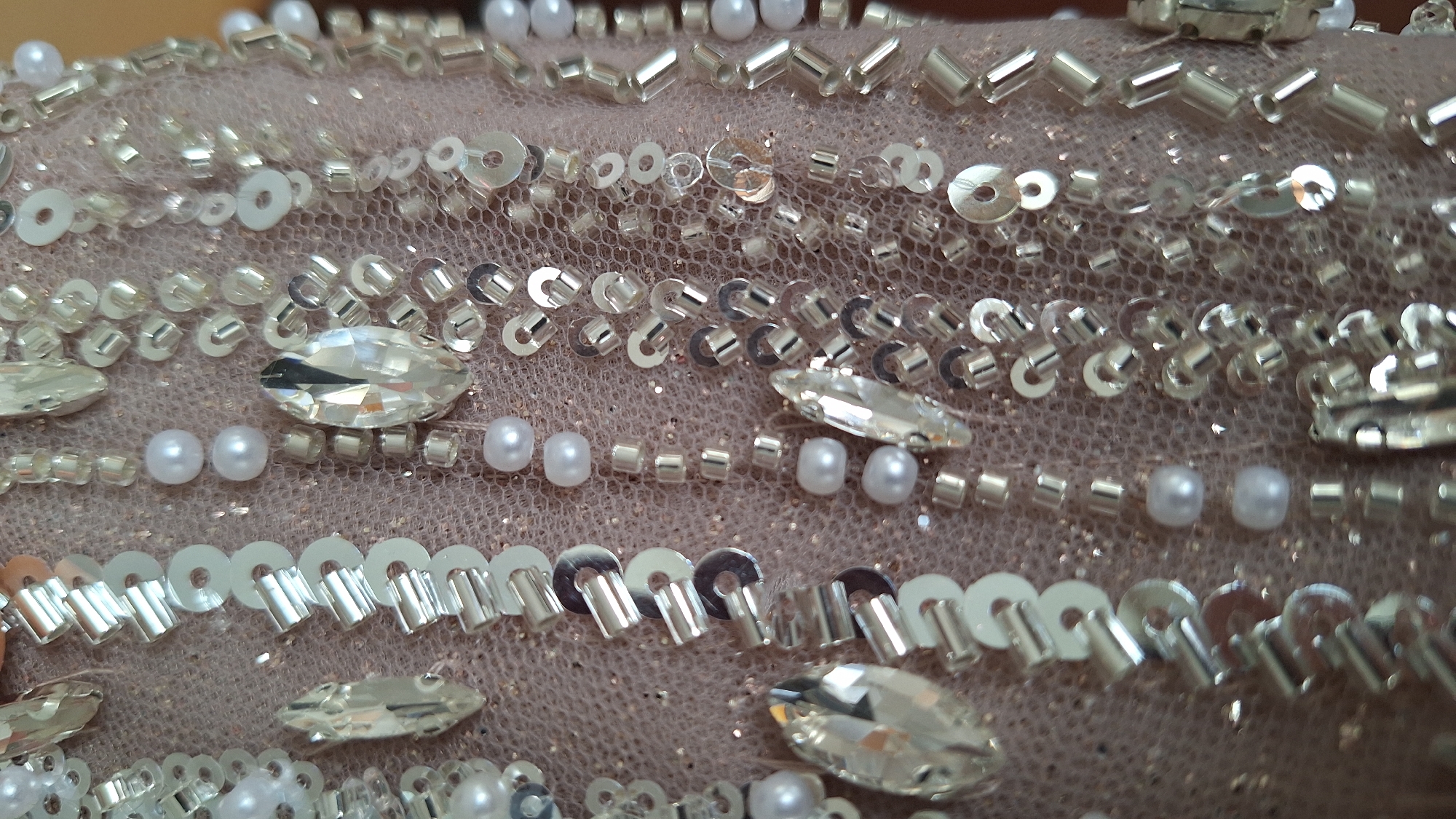 The image shows a close-up of fabric with intricate beadwork and embellishments. The fabric appears to be a sheer, possibly nude-colored material, overlaid with a dense arrangement of sequins, beads, faux pearls, and large, faceted crystals resembling rhinestones. The beads come in various shapes and sizes, including cylindrical beads and round beads. The sequins are small and disc-shaped, creating a shimmering effect. The embellishments are arranged in rows and patterns, giving the fabric a luxurious and decorative appearance.
There is no information in the provided image that indicates the location to be Blagoevgrad, Bulgaria.
