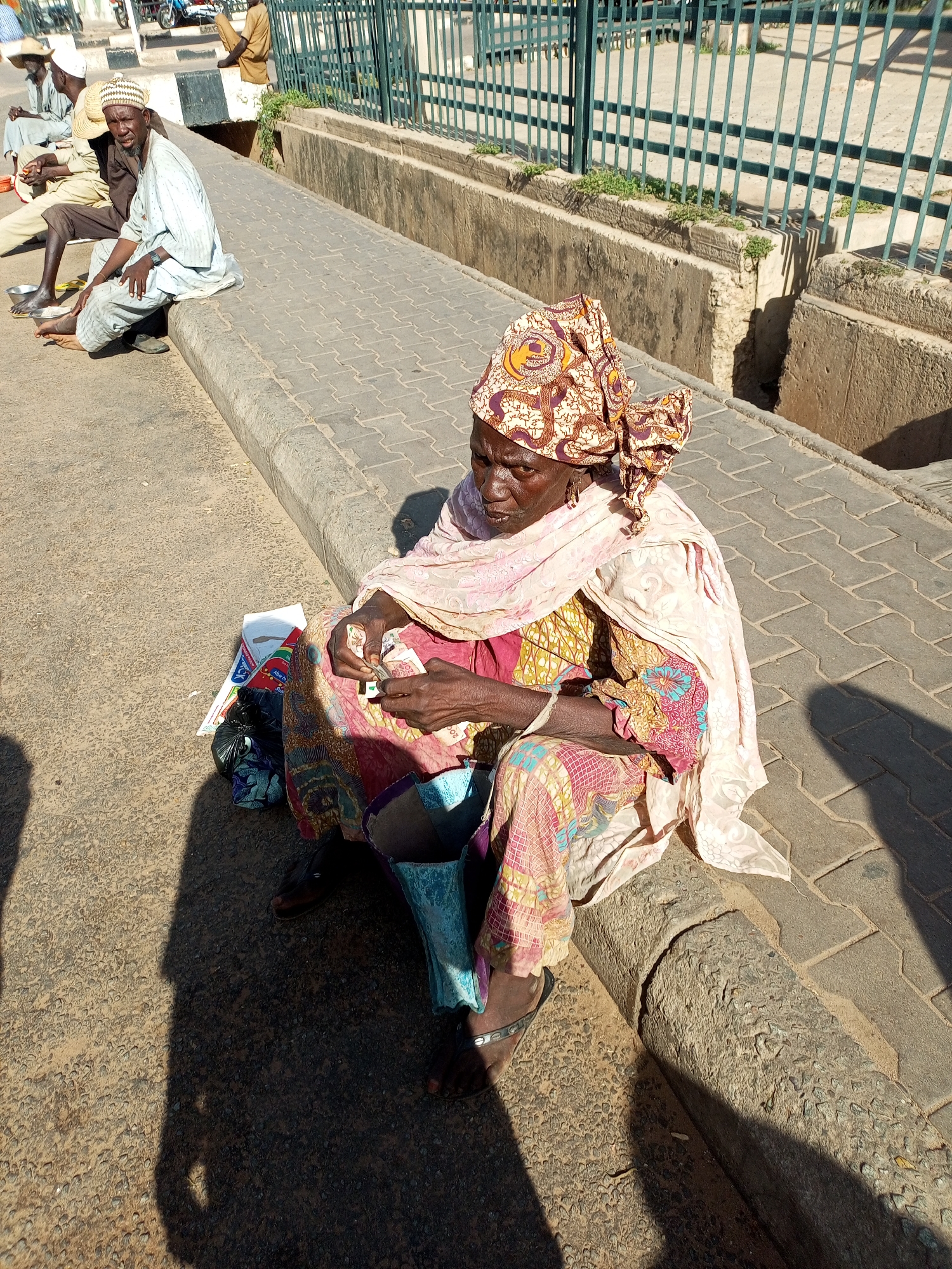 The image depicts a scene on a sidewalk in Arawa, Nigeria. In the foreground, an elderly woman is seated on the curb, wearing a colorful headscarf and patterned clothing. Her hands are occupied with holding and counting banknotes, suggesting an interaction involving money. To her left, a pile of assorted bags and what appears to be a newspaper or magazine are visible.
Further back on the sidewalk, several men are also seated. One man, wearing a light-colored robe and a straw hat, is looking towards the camera. Another man, in a darker robe and a white cap, sits beside him. Behind them, a man in a light-colored robe and a hat is seated further down the sidewalk. In the background, a green metal fence encloses a grassy area and a concrete structure. The ground surface is paved with interlocking bricks on the sidewalk and asphalt on the road. The lighting suggests it is daytime, with shadows cast across the scene.
