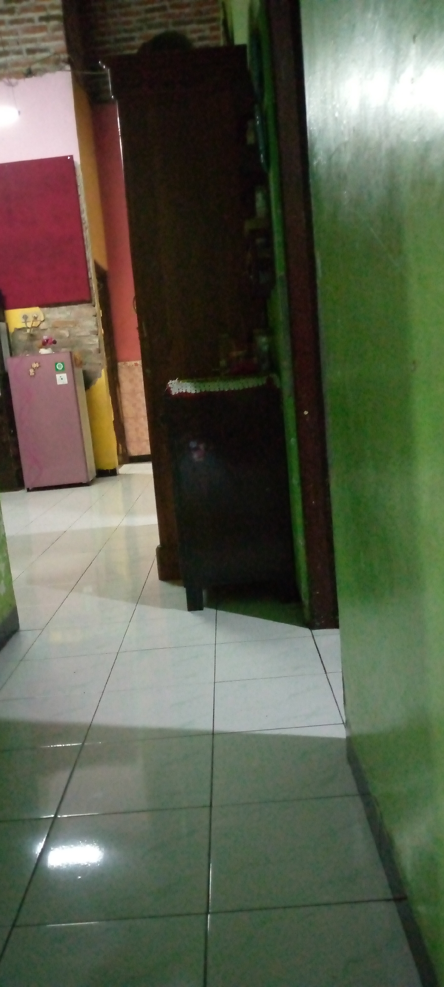 This image shows an indoor scene, possibly a residential hallway or kitchen area, in Sumberwono, Indonesia. The floor is tiled with white square tiles. To the left, a pink refrigerator stands against a wall. The wall to the left of the refrigerator is red, and behind it, a brick wall is visible. A yellow object, possibly a curtain or a rolled-up mat, is leaning against the wall next to the refrigerator. In the center of the image, a tall, dark brown wooden cabinet or wardrobe is present. A portion of a green wall is visible on the right side of the frame. The scene appears to be dimly lit, with reflections on the tiled floor suggesting a light source from above or the side.