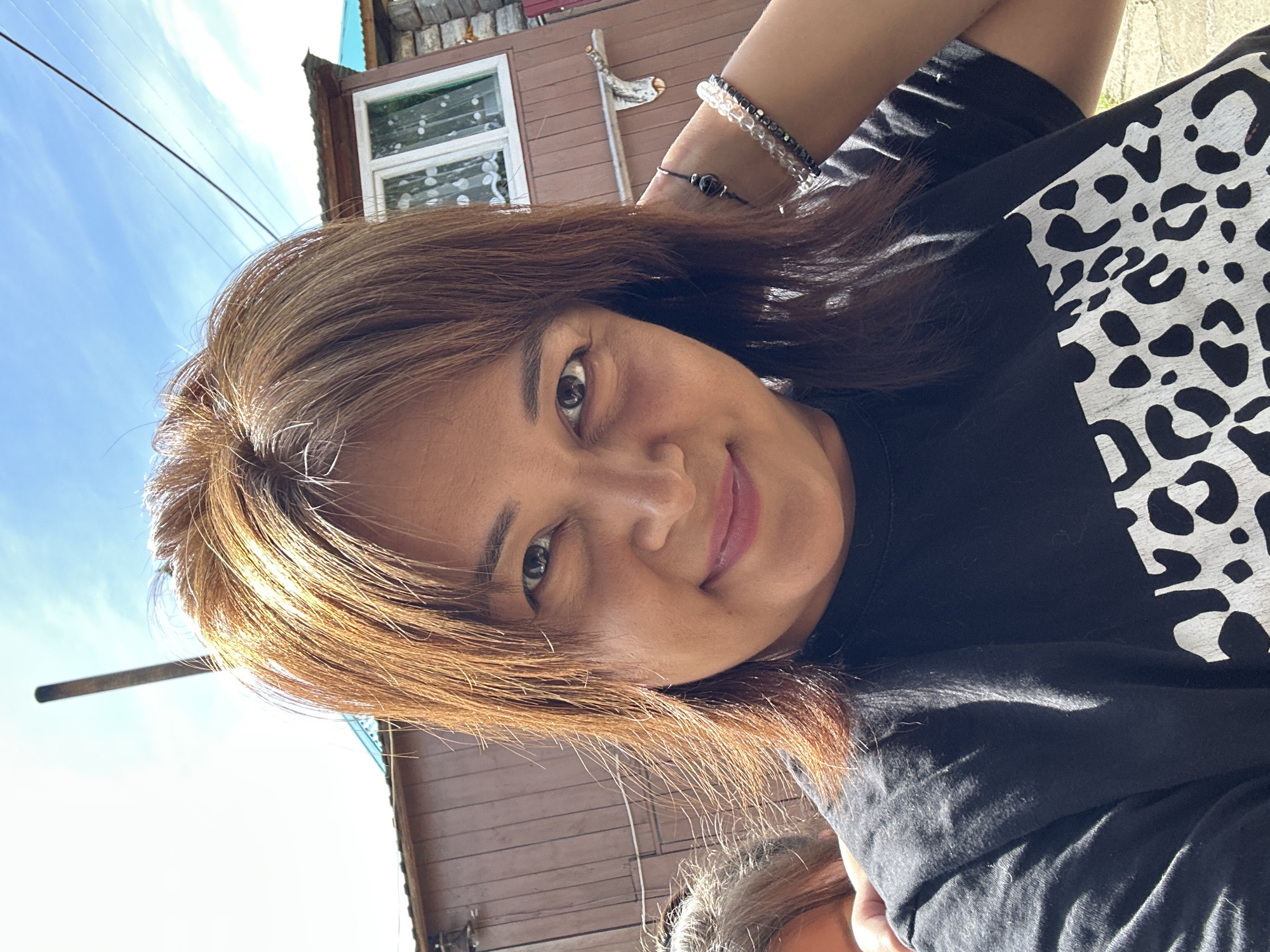 A woman, with long auburn hair and brown eyes, is captured in a close-up selfie. She is wearing a black t-shirt with a white leopard print design on the right side of her chest. Around her left wrist, she wears two beaded bracelets. Her gaze is directed towards the camera, and her lips are slightly parted, suggesting a gentle smile or relaxed expression.
The background indicates an outdoor setting, with a glimpse of a wooden building and a bright blue sky with thin white clouds. Power lines are visible against the sky. The lighting suggests it is daytime, with sunlight casting shadows and highlighting her hair. The overall impression is a casual, everyday moment. There is no visible text in the image.